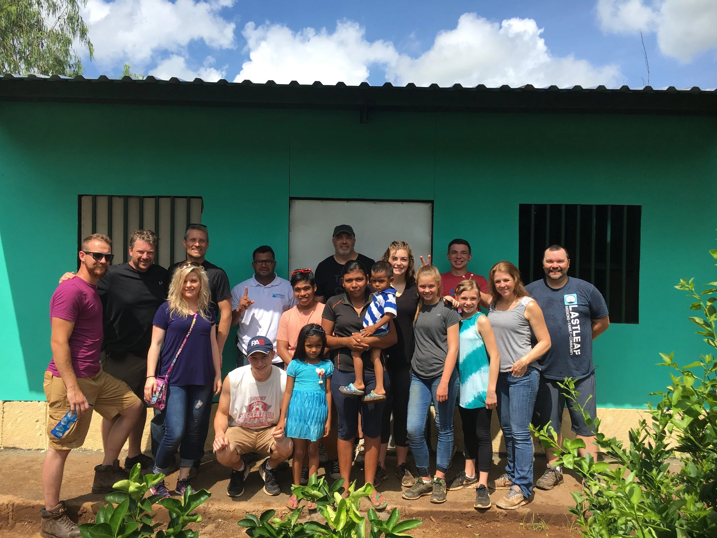 Group of adults and children standing outside in front of a turquoise building with windows and a door, under a partly cloudy sky.