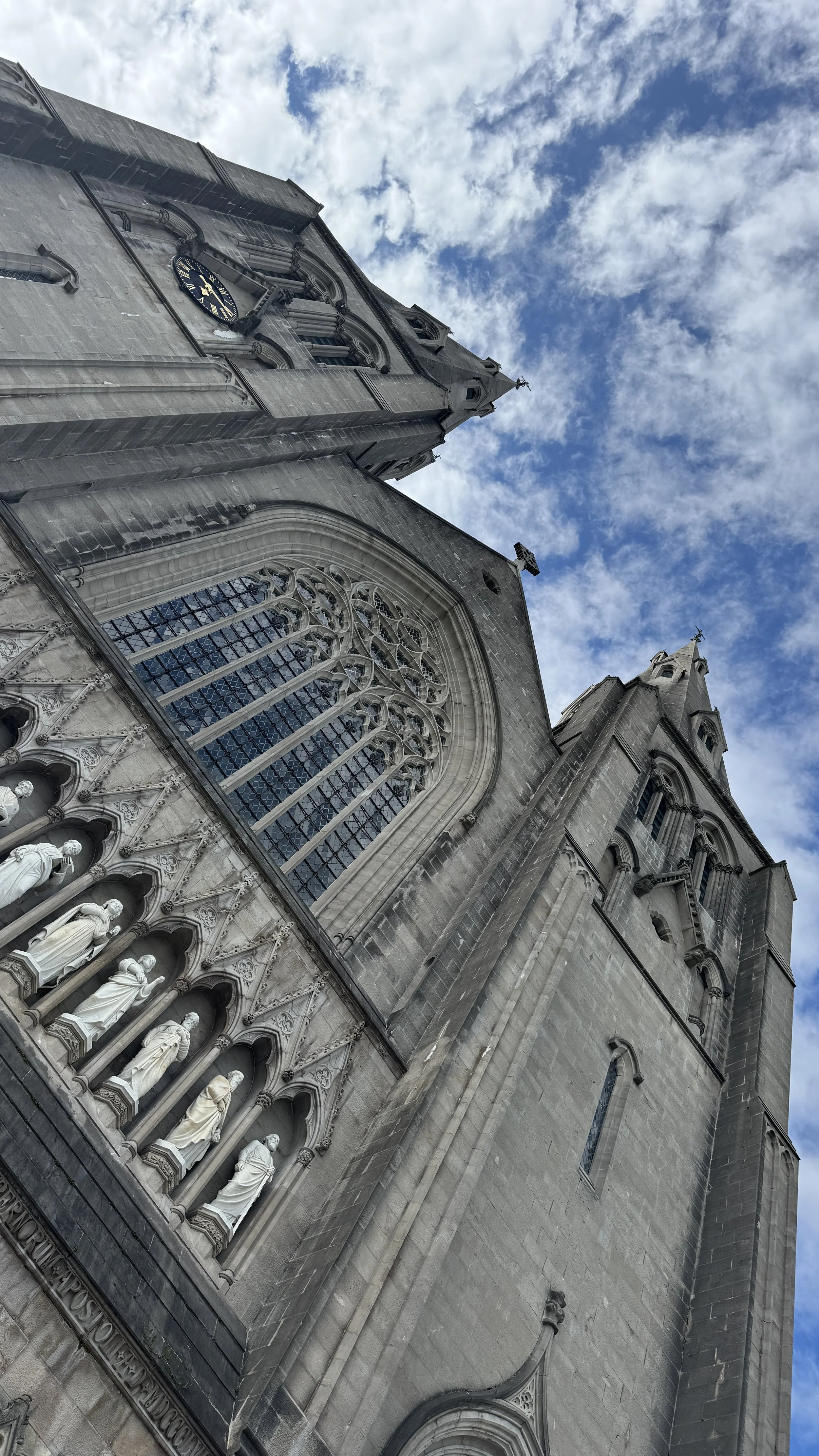 Looking up at a tall, gray stone Gothic church with statues, a large stained-glass window, and a clock on the tower, against a partly cloudy sky.