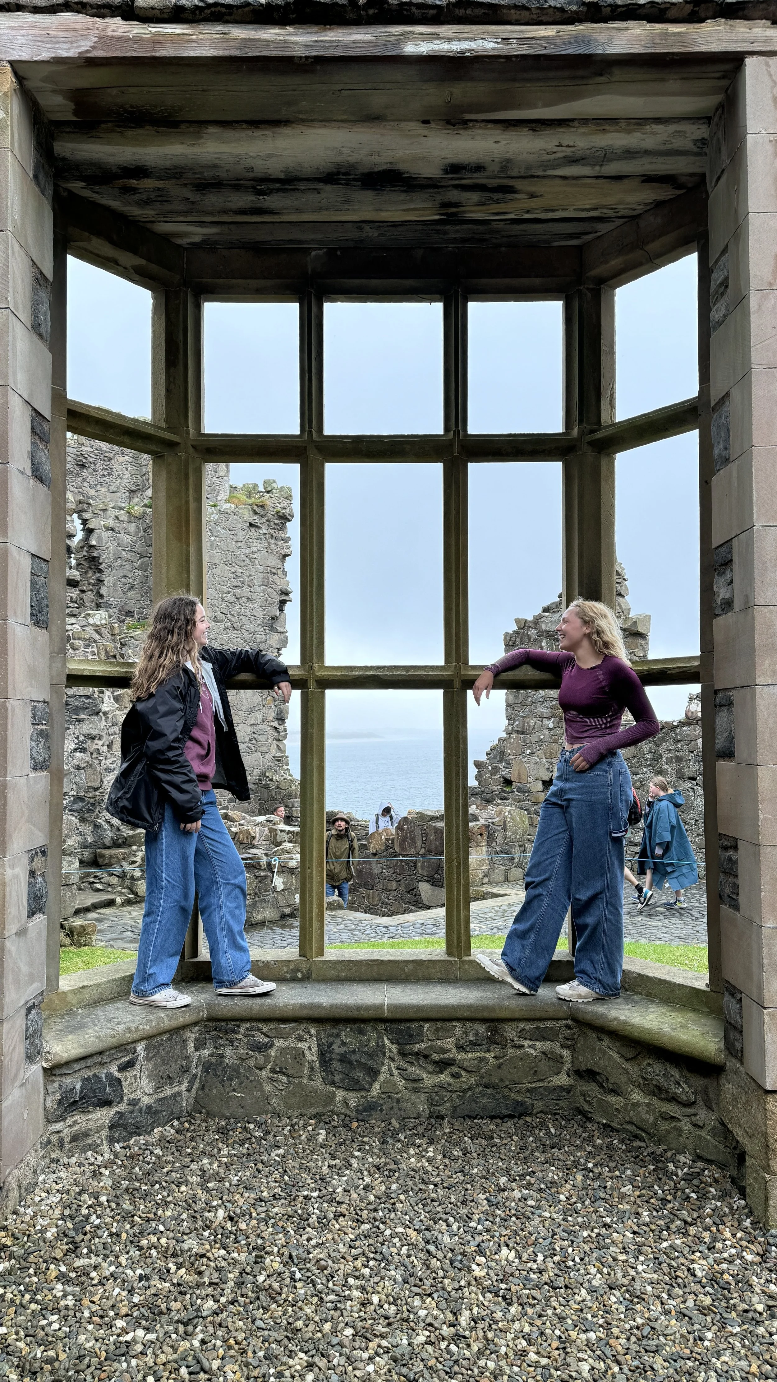 Two young women with long hair standing and leaning on a window frame at a historic stone site, facing each other, with other visitors in the background overlooking a rocky coastline and ocean, under an overcast sky.
