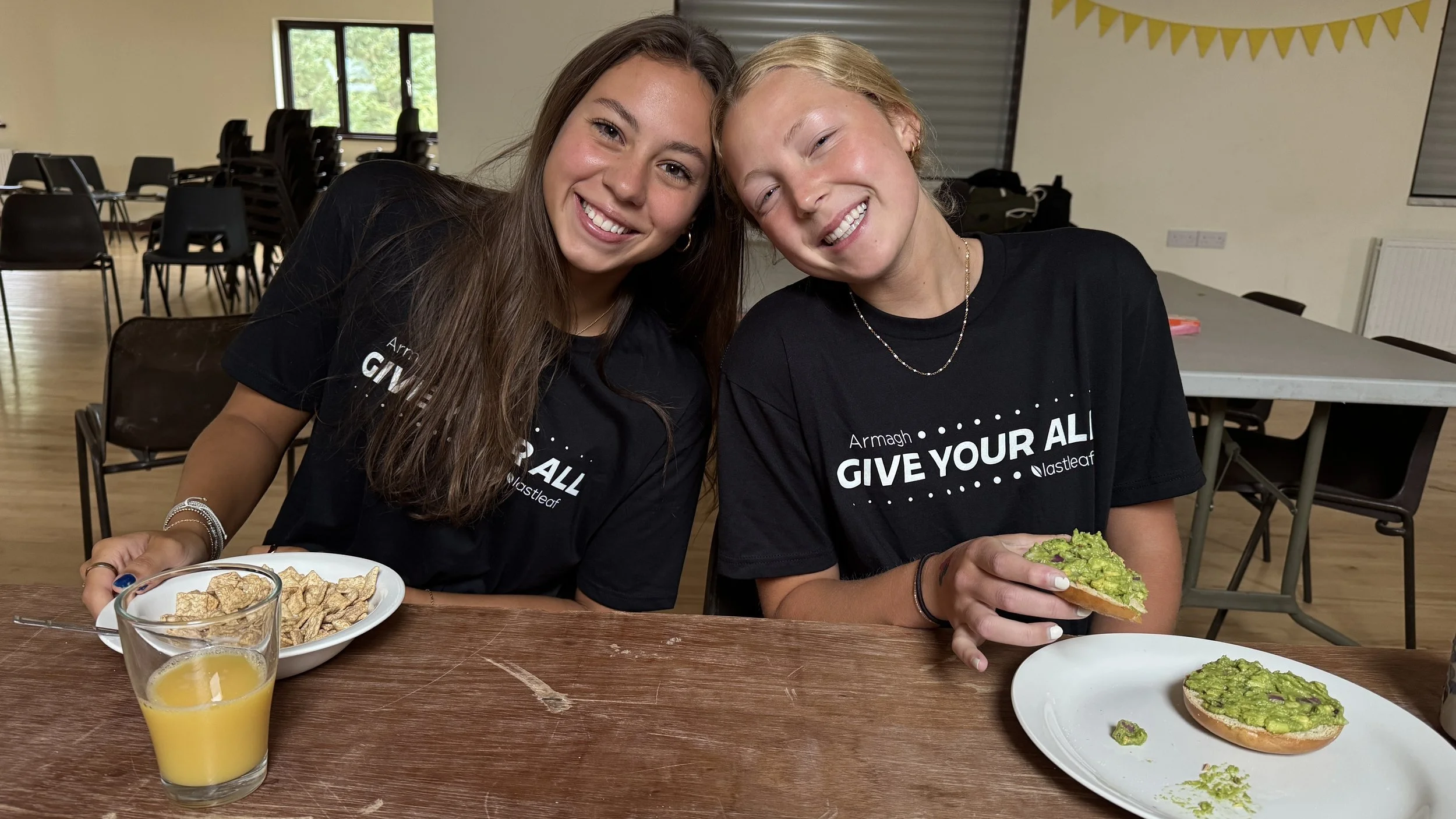 Two young women sitting at a table, smiling, wearing matching black t-shirts that say 'Give Your All.' One woman has long brown hair and the other has blonde hair. They are holding plates with avocado toast, and there is a glass of orange juice on the table. In the background, there are chairs and tables in a large room.