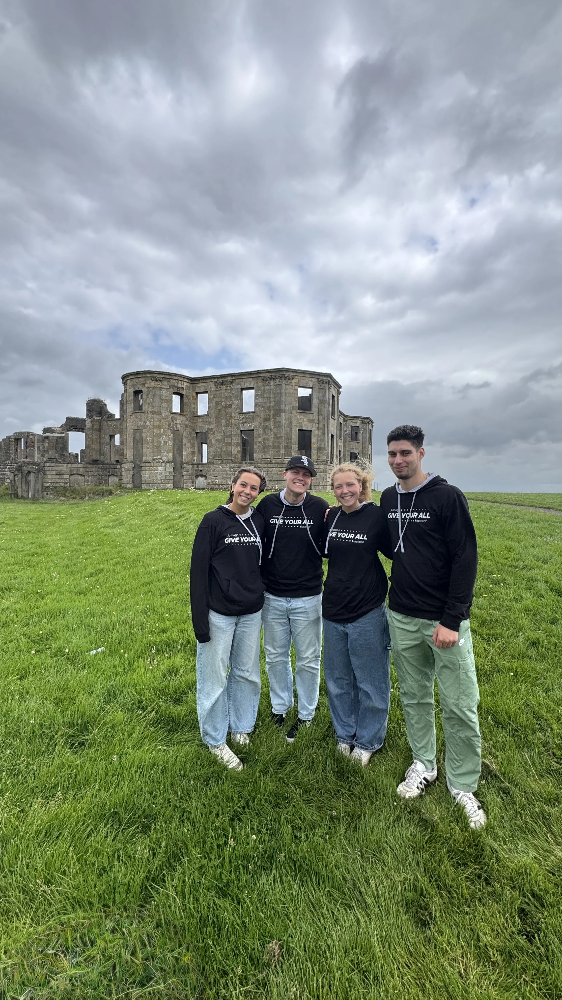 Four young adults standing on a grassy field in front of a historic stone building with a cloudy sky above. They are smiling and wearing matching black hoodies.