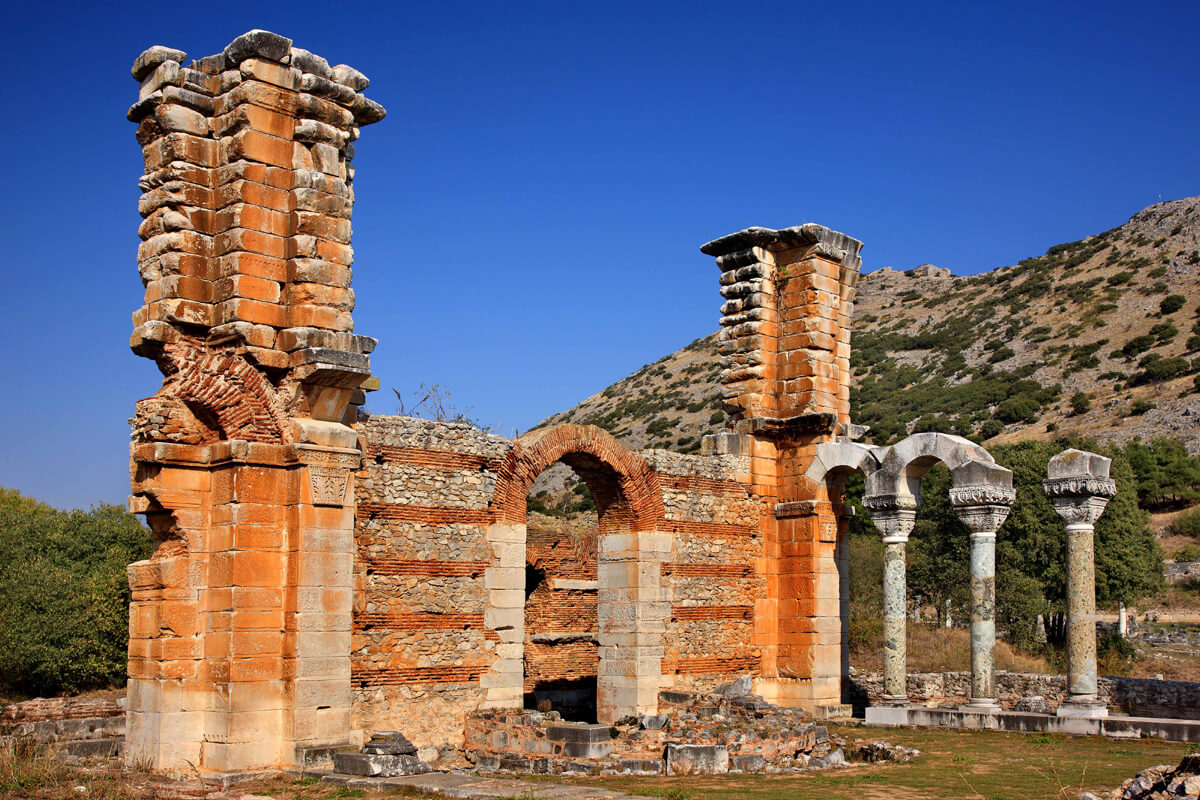 Ancient ruins of a stone and brick structure with columns and an arch, set against a mountainous landscape and clear blue sky.