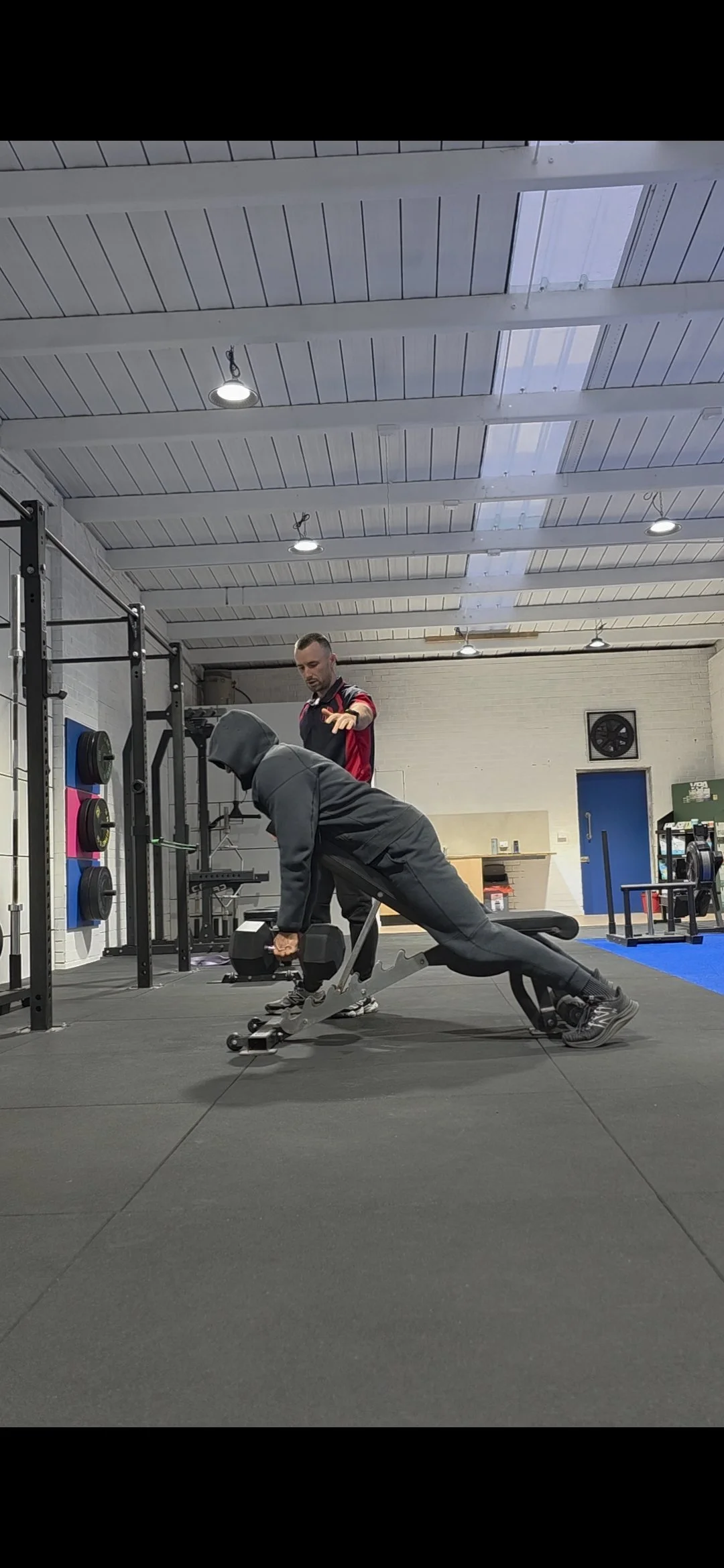 A person in a hoodie performing a plank exercise with a dumbbell in a gym, guided by a trainer.