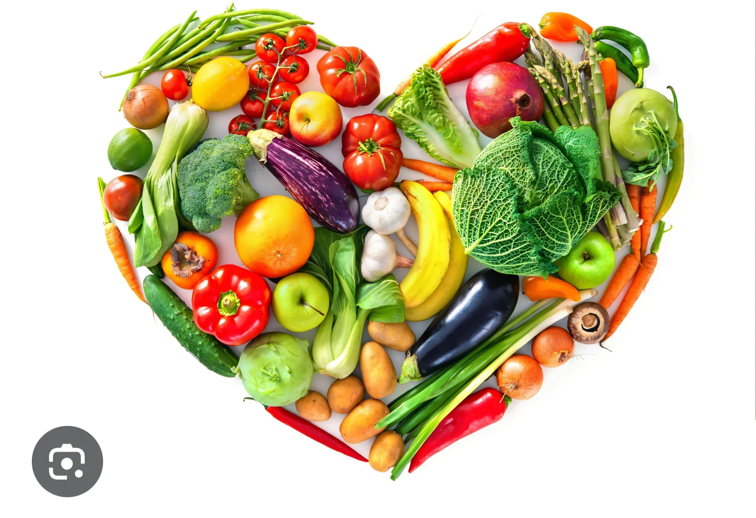 A heart-shaped arrangement of various fresh vegetables and fruits including tomatoes, bell peppers, cucumbers, zucchinis, carrots, garlic, onions, bananas, eggplant, lettuce, and other produce set against a white background.