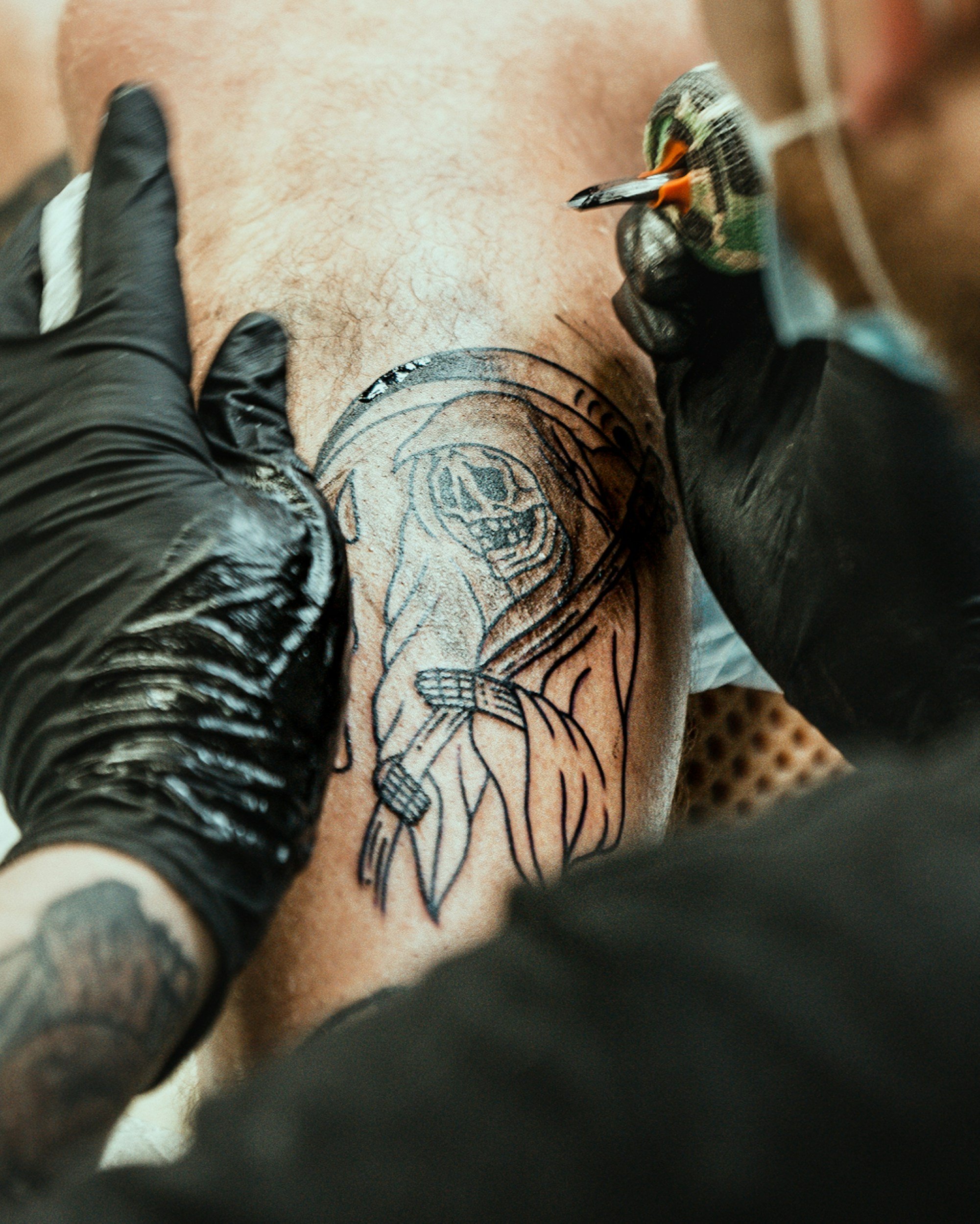 A tattoo artist is creating a tattoo of a skeleton reading a book on a person's arm, using a tattoo machine while wearing black gloves.