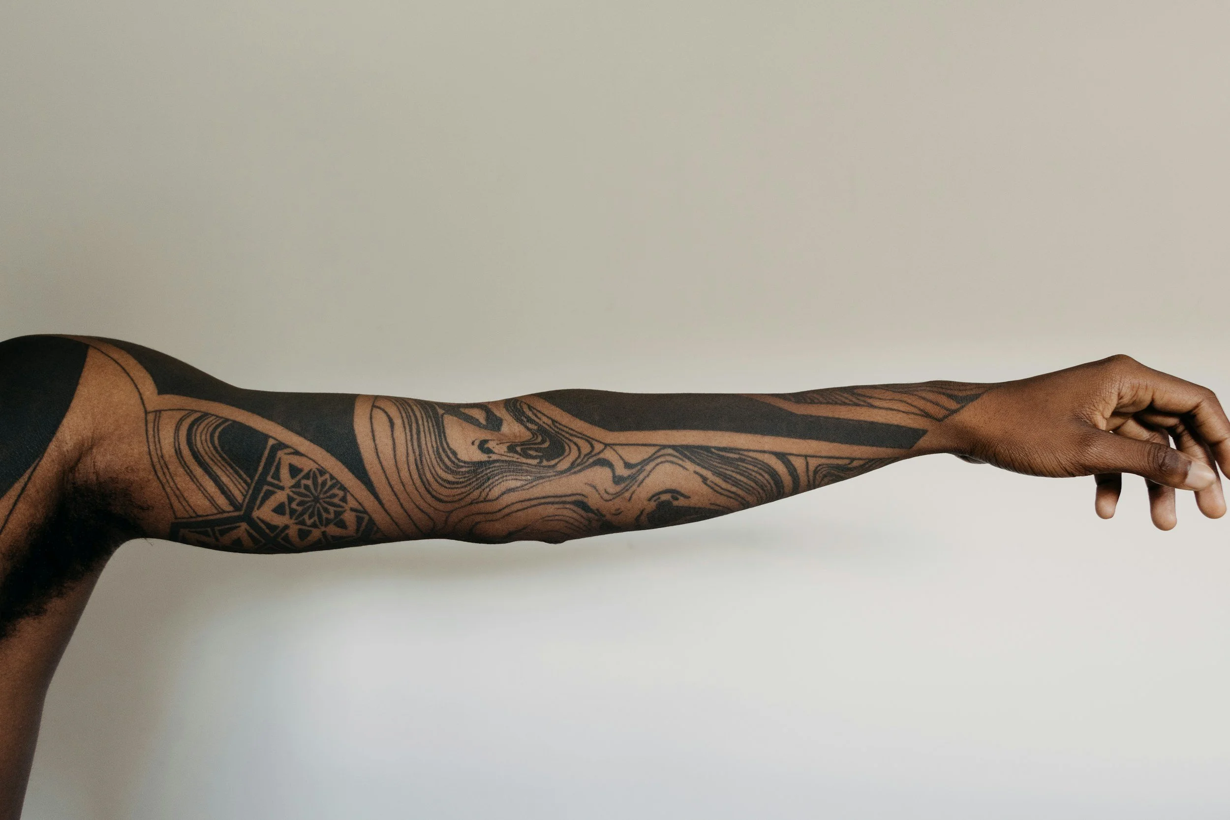 A person's outstretched arm with intricate black tattoos covering the arm, hand, and part of the shoulder, set against a plain light-colored background.