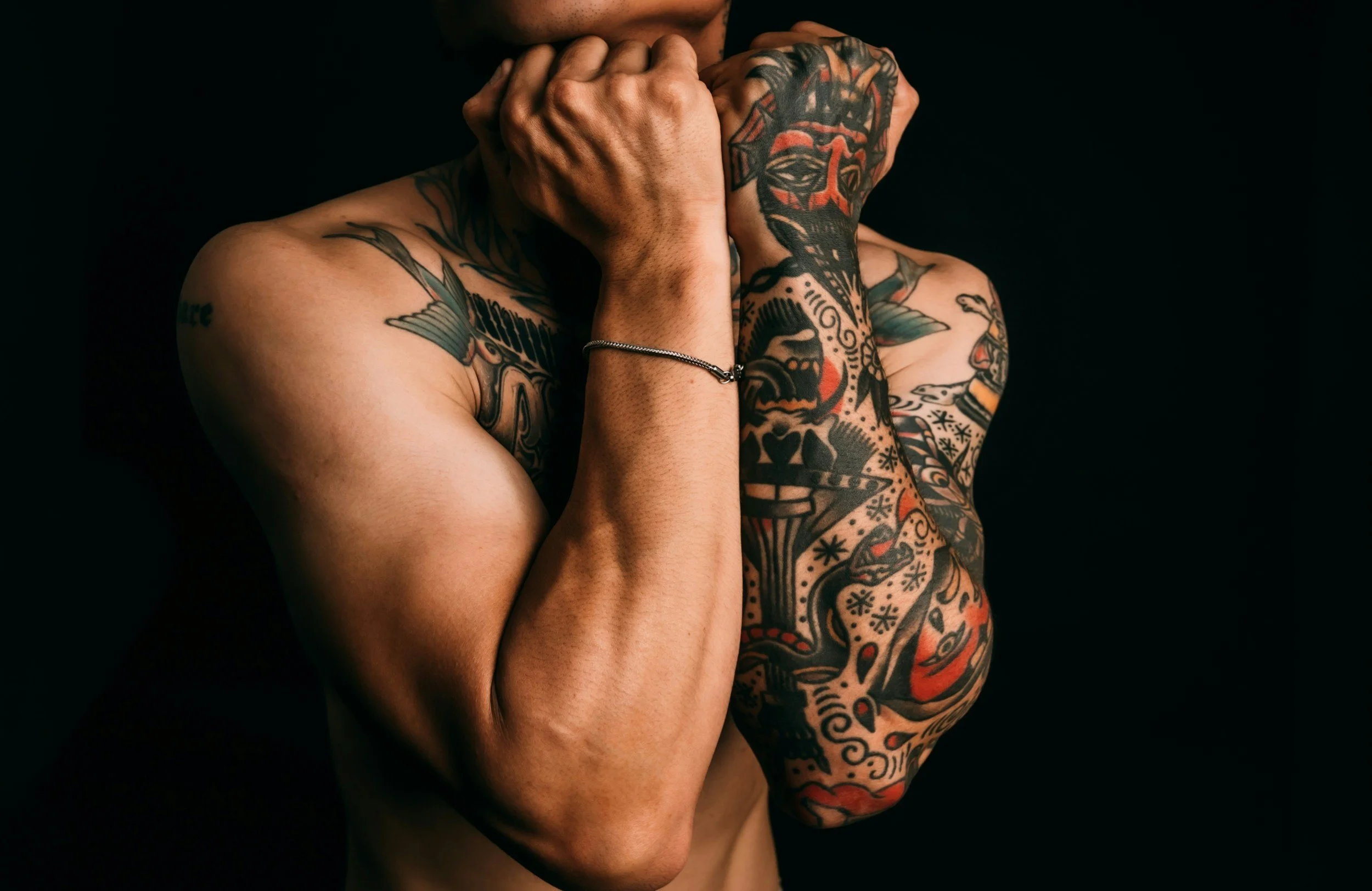 Person with tattoos covering their arms, shoulders, and chest, covering their face with their hands, wearing a bracelet, against a dark background.