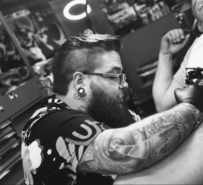 A man with glasses, tattoos, and a beard sitting at a tattoo station, receiving a tattoo on his arm from an artist.