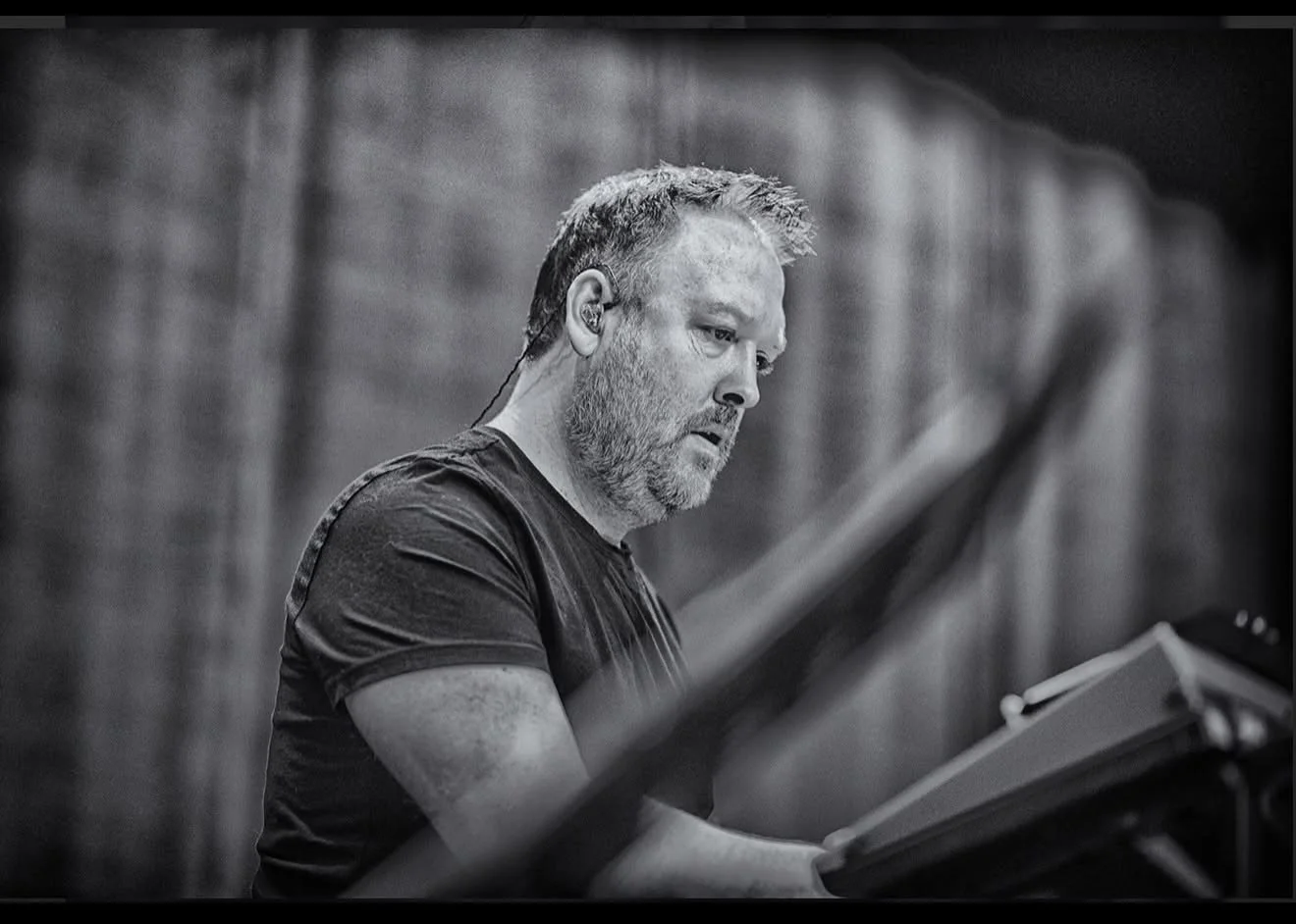 A man with short hair and a beard wearing a black T-shirt, working at a keyboard with a focused expression.