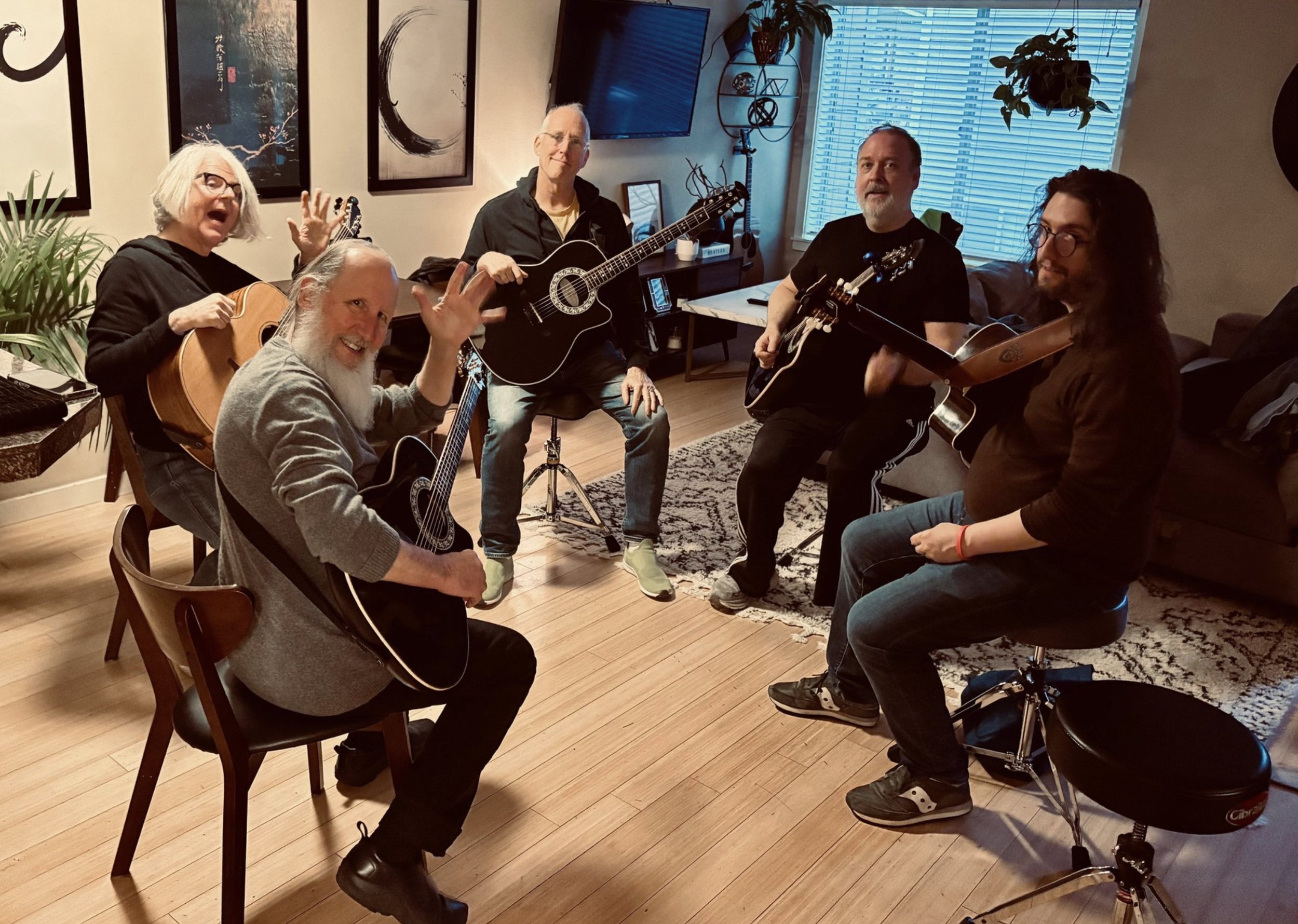 Five people with guitars in a cozy living room, four sitting and one standing, appear to be preparing to play or practicing music together.