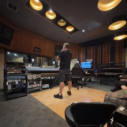 Two people in a recording studio, one standing and one sitting at a computer workstation, surrounded by audio equipment and soundproof walls.