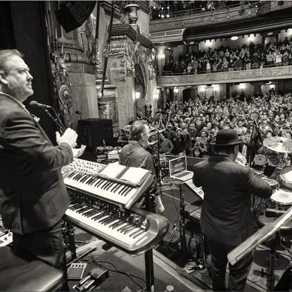 Musicians performing on stage in front of a large audience in a theater with ornate balconies and decor.