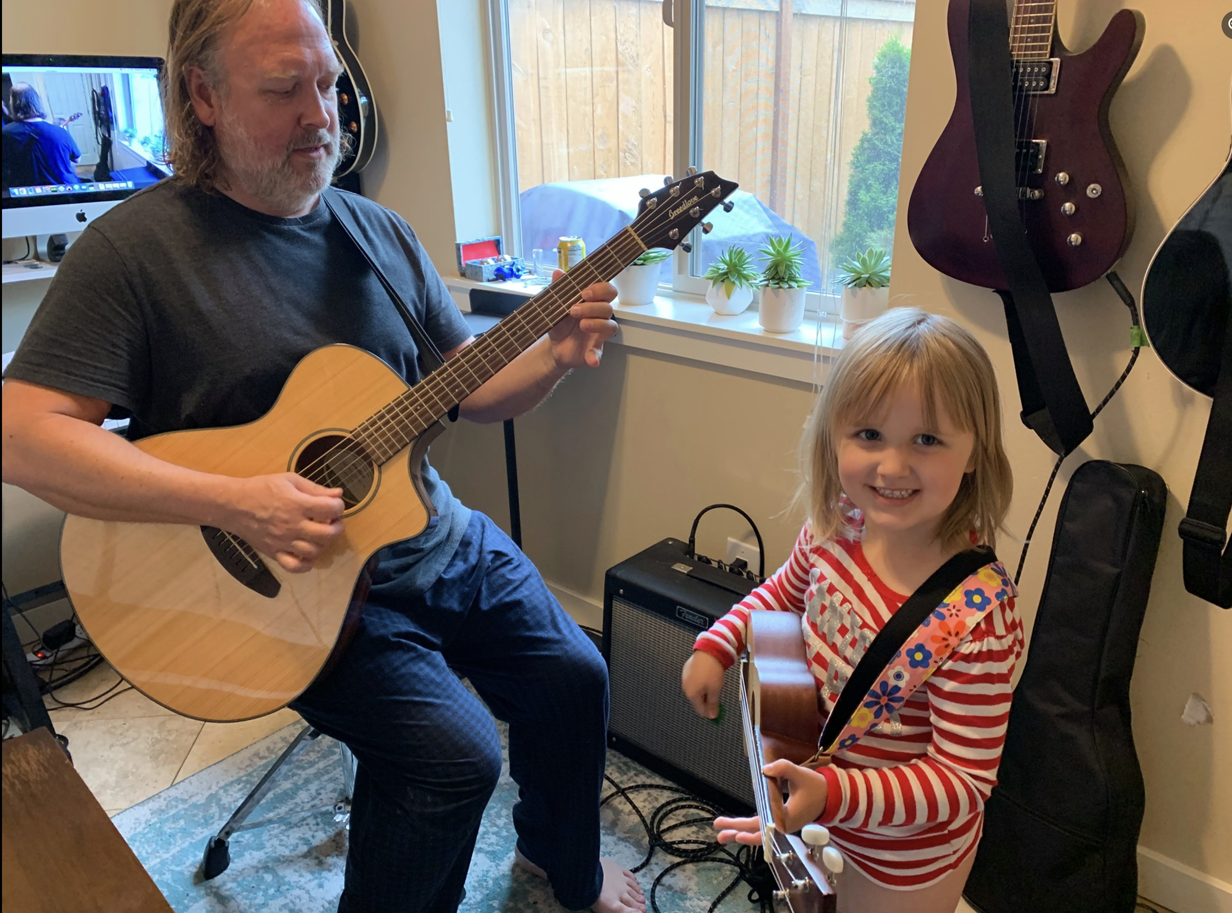 A bearded man playing an acoustic guitar while sitting on a chair, and a young girl in a red and white striped shirt smiling and holding a guitar, in a room with guitars and a small amplifier.