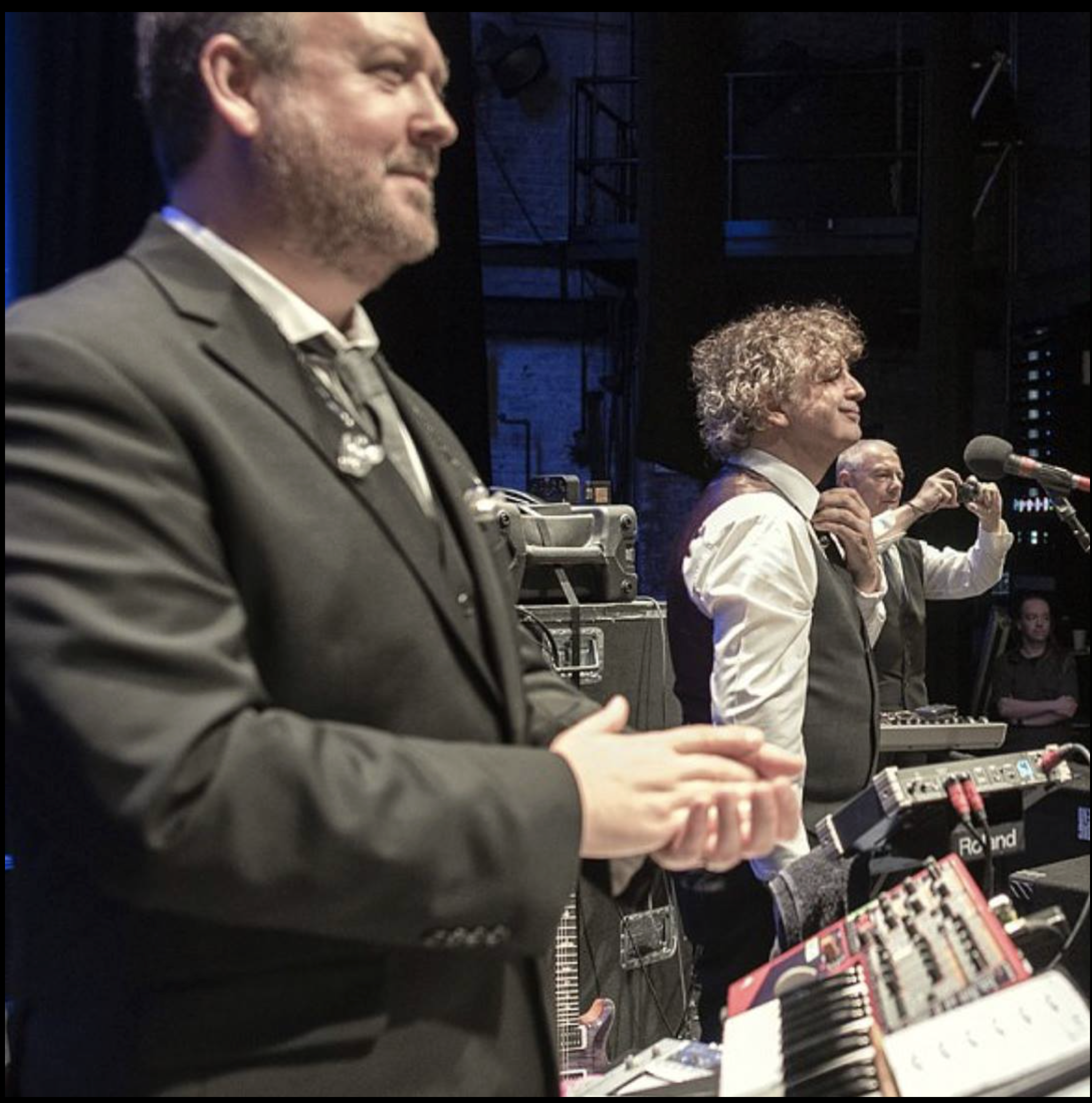 Three musicians in formal attire perform on stage with musical equipment.