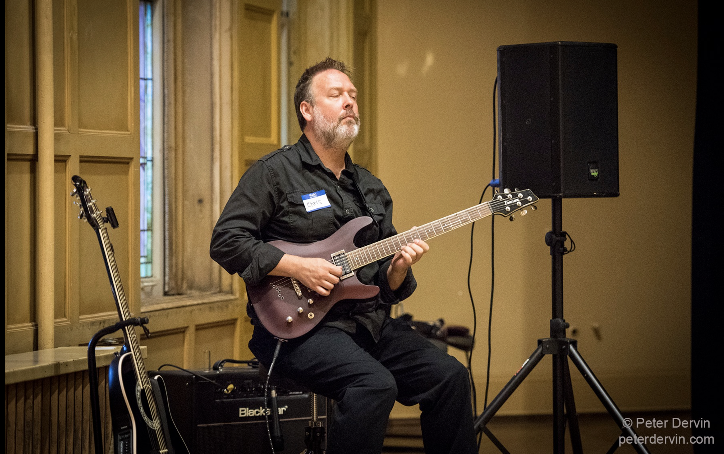 A man with a beard and dark hair playing an electric guitar while seated. He is wearing a black shirt with a name tag. Behind him, there is an acoustic guitar leaning against an amplifier. The setting appears to be an indoor space with wooden paneling and a window.