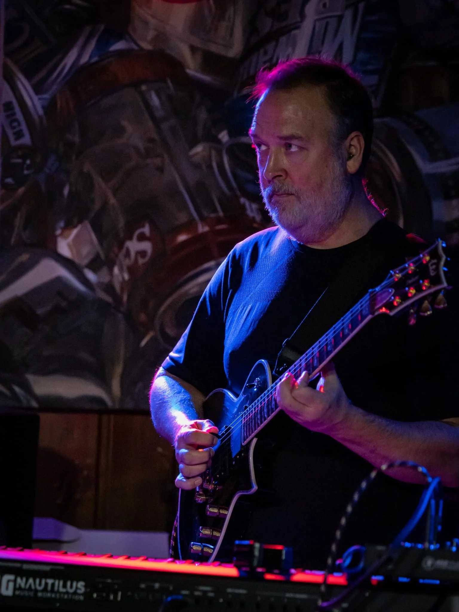 A man with gray hair and beard playing electric guitar in a dimly lit room with stacked drum cases in the background.