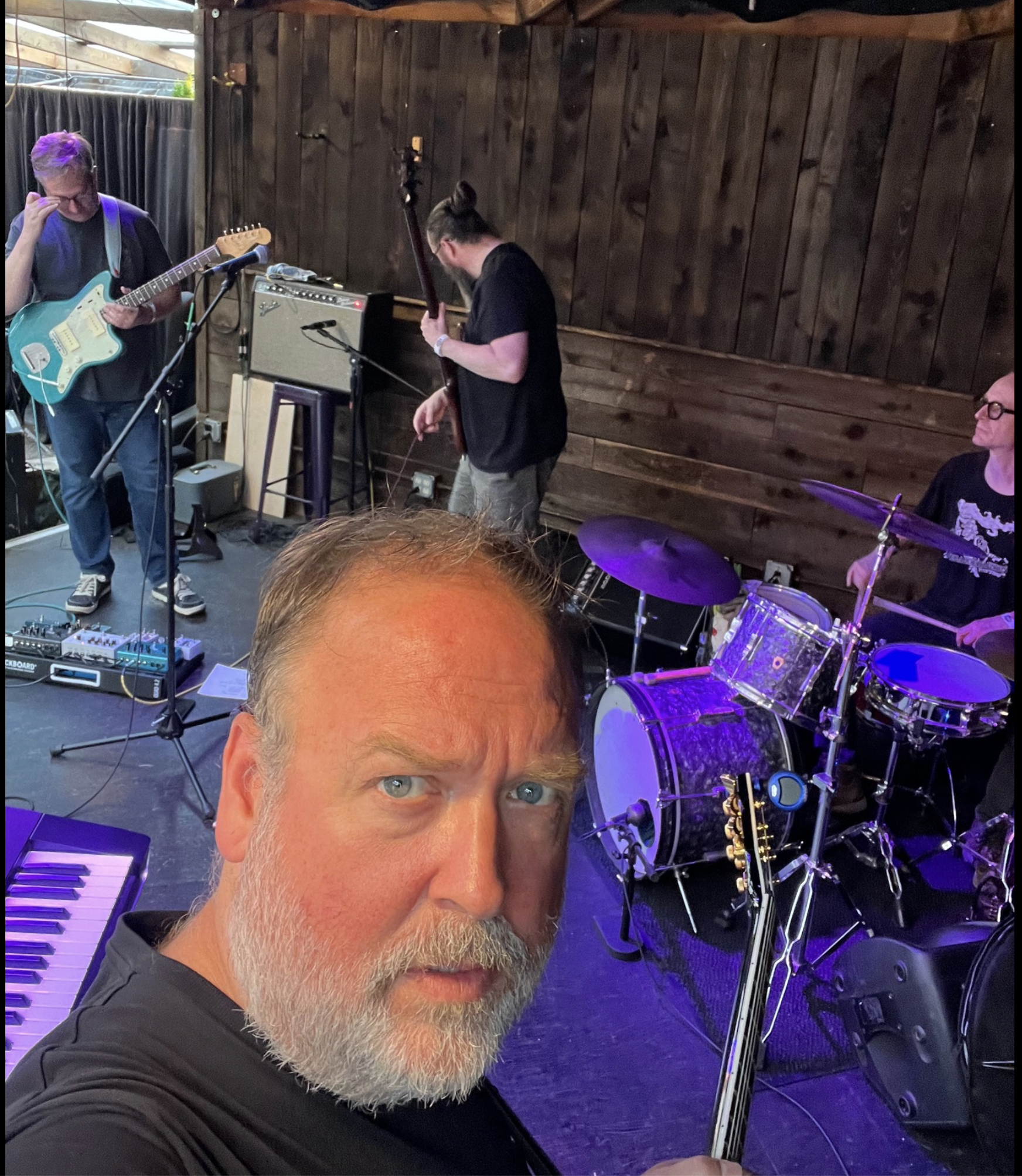 Man with a gray beard taking a selfie in front of a band performing on a stage with wooden walls, including a guitarist, a bassist, and a drummer.