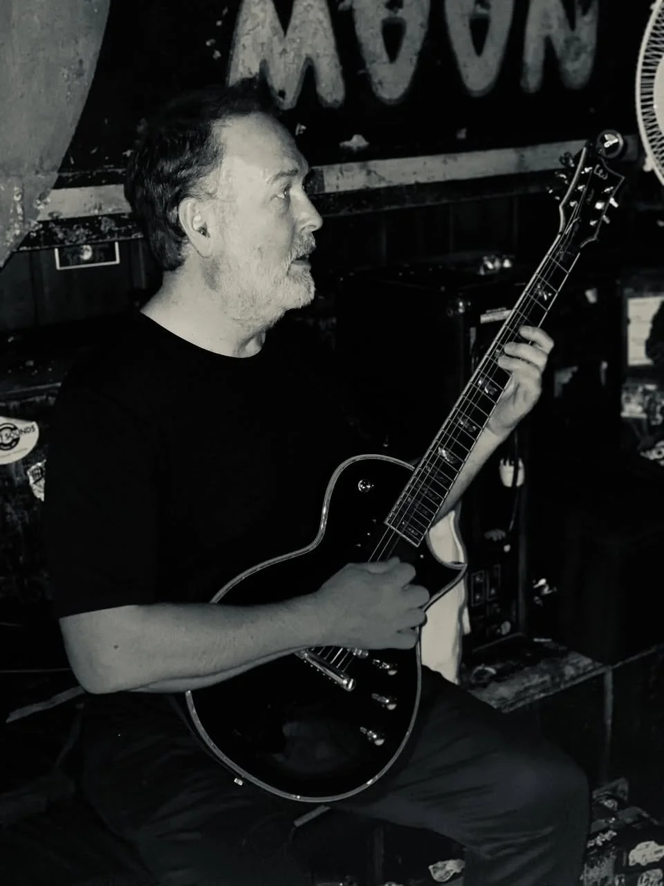 A middle-aged man with gray hair and a beard playing an acoustic guitar in a dimly lit bar or music venue.