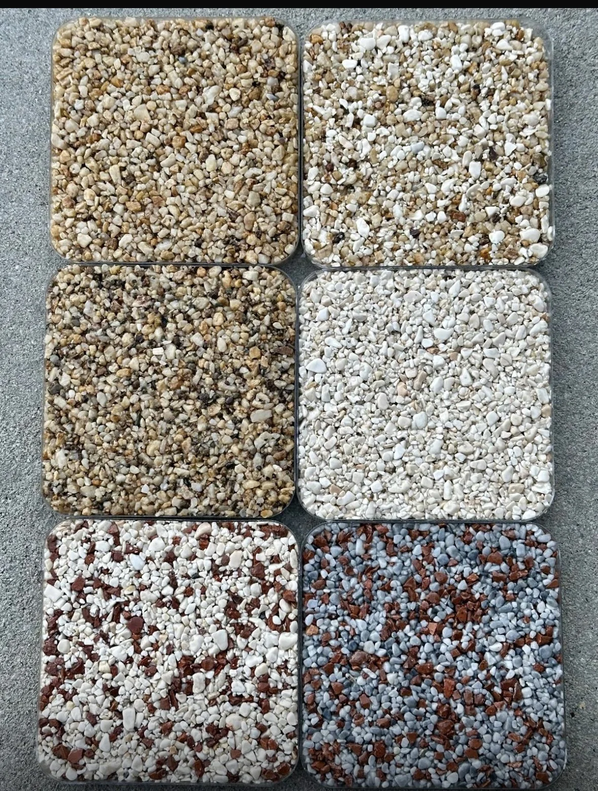 Six square trays filled with various small pebbles and gravel, arranged in two rows of three on a concrete surface.