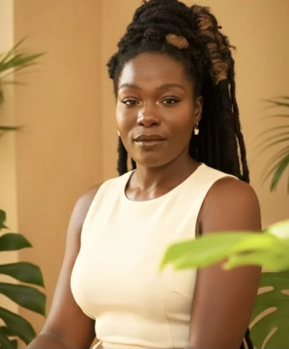 A woman with dark skin, natural dreadlocks with blonde highlights, wearing a cream sleeveless top, standing indoors with green plants in the background.