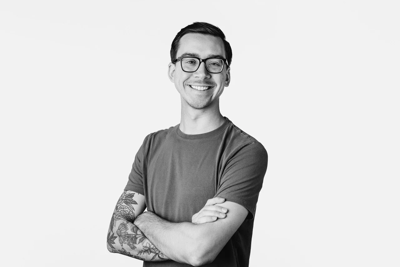 Black and white photo of a smiling man with dark hair, wearing glasses and a casual t-shirt, posing with arms crossed against a plain light background.