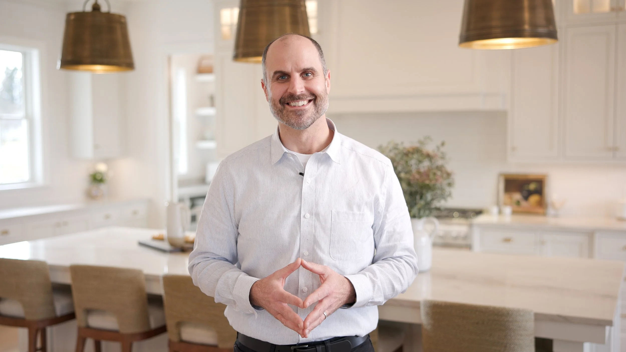 Smiling man in white shirt standing in a bright kitchen with white cabinets, large pendant lights, and a dining table.