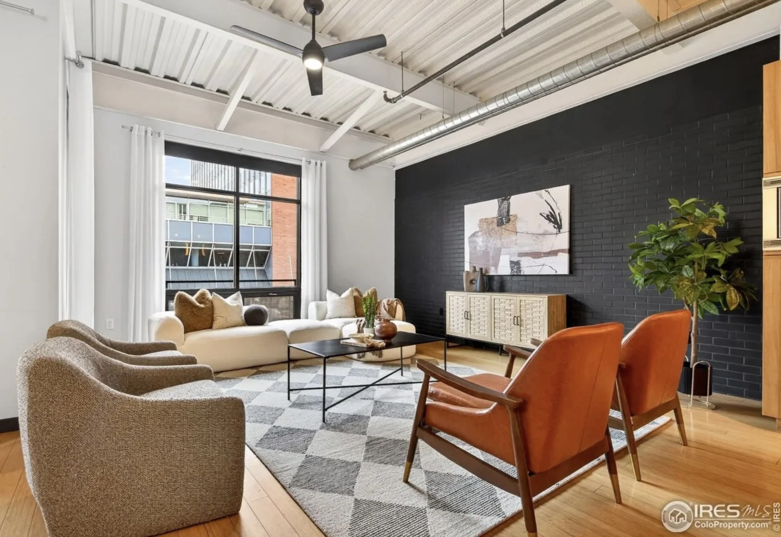 Modern living room with white walls, large window with white curtains, a black accent wall, wooden flooring, six chairs including a white sofa and orange armchairs, a black coffee table, a white textured sideboard, framed abstract art, and a potted plant.