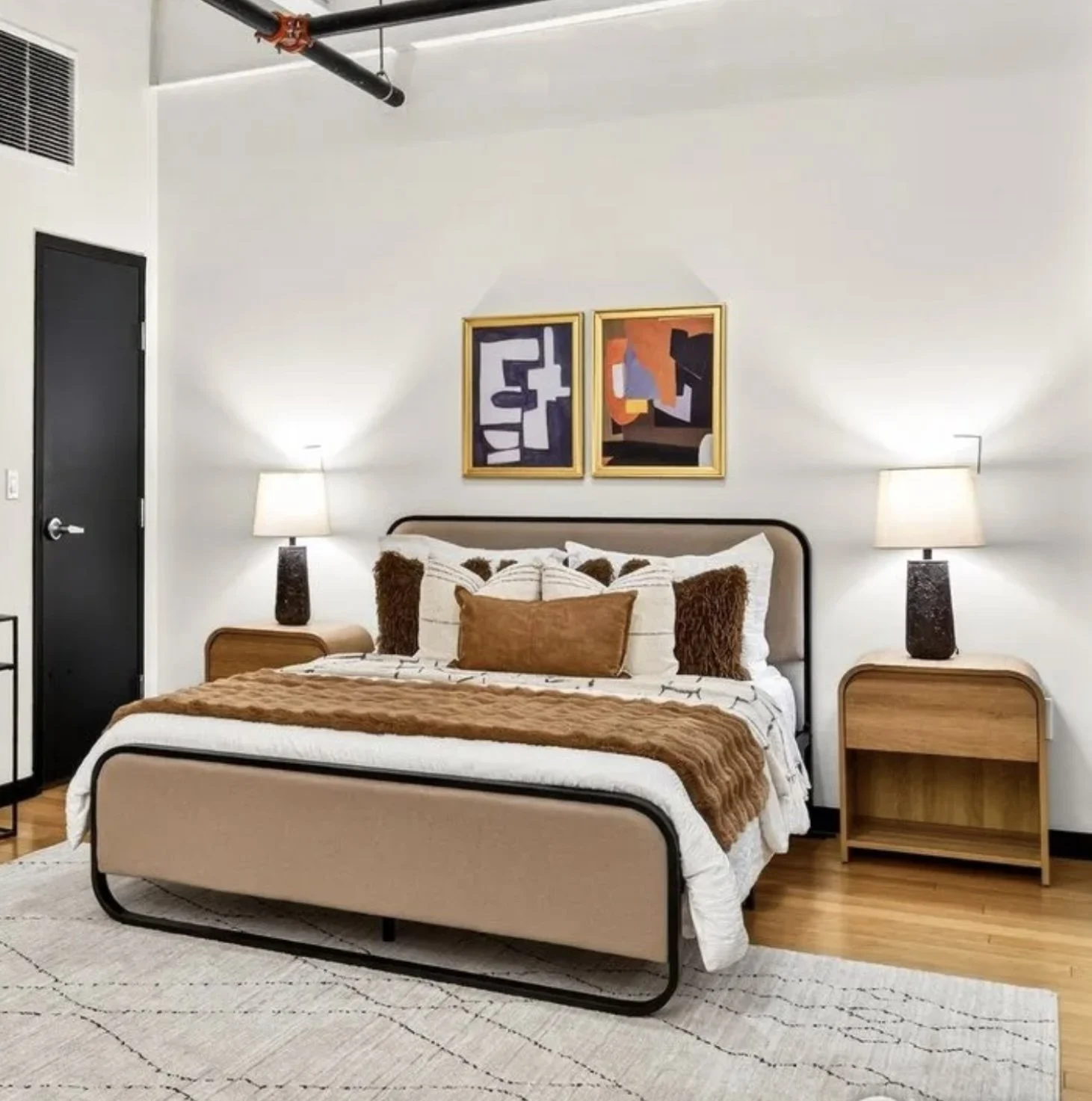 A neatly made bed with white sheets, brown and white pillows, and a brown throw blanket in a modern bedroom. There are matching nightstands with lamps on each side, abstract paintings above the bed, and hardwood flooring with a light-colored area rug.