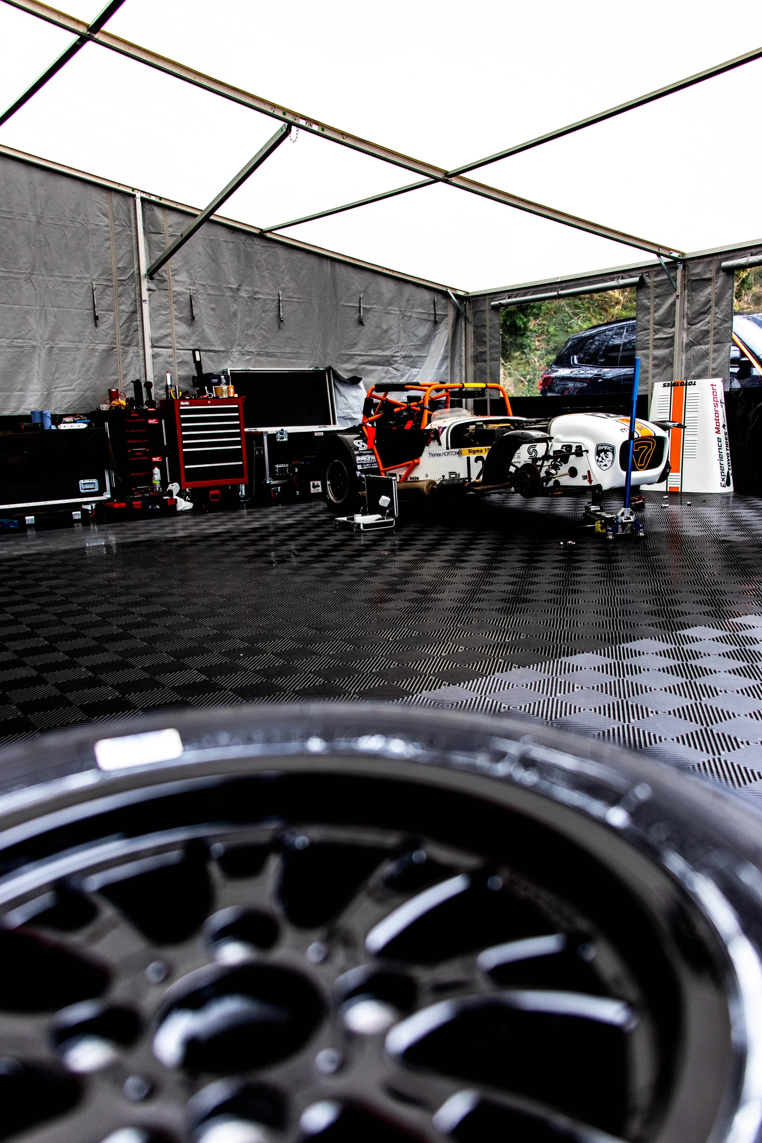 View from the back of a race car, looking through the wheel wells at a race car in a garage, with tools and equipment nearby.