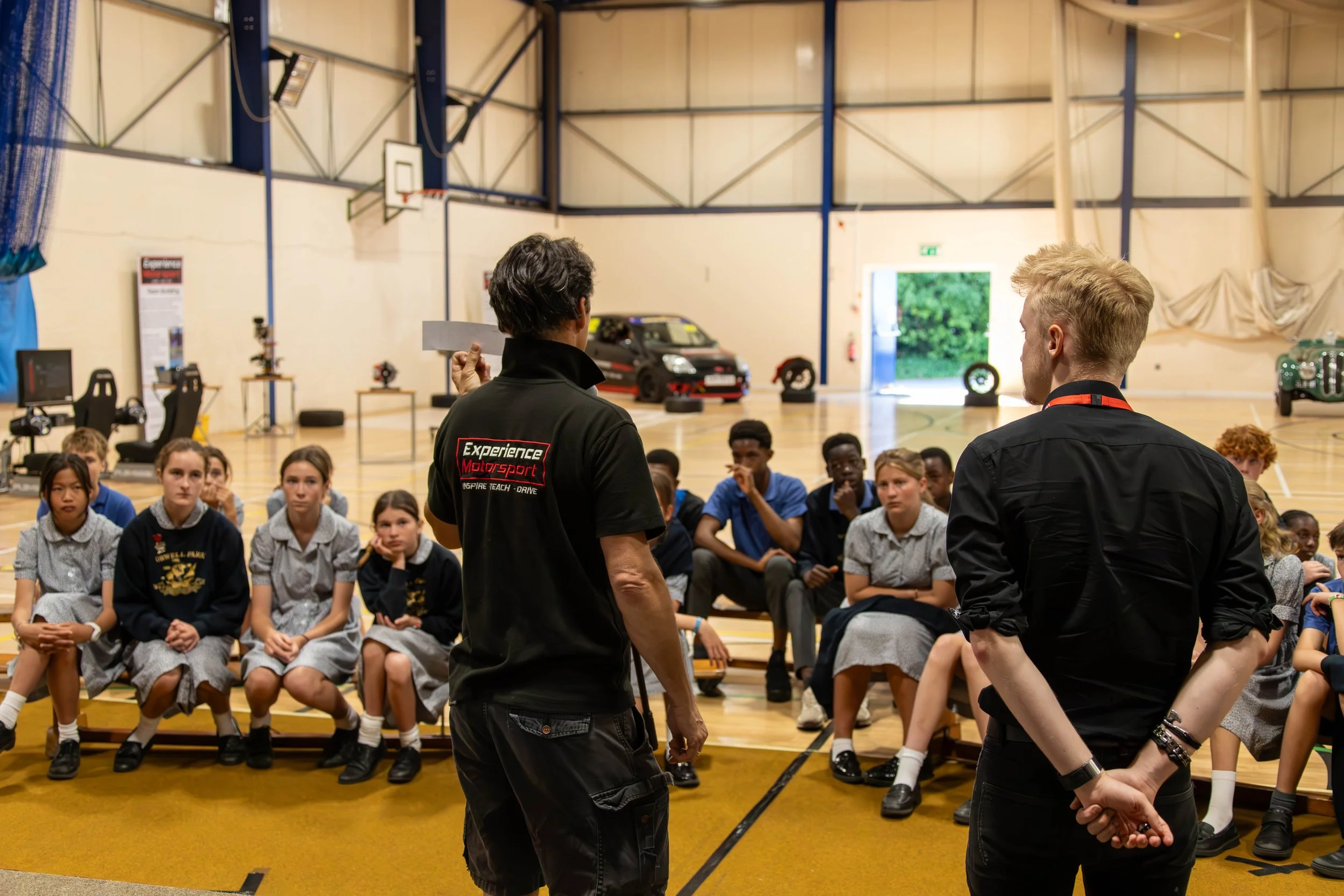 A group of school children sitting on chairs in a gymnasium, listening to the Experience Motorsport team.