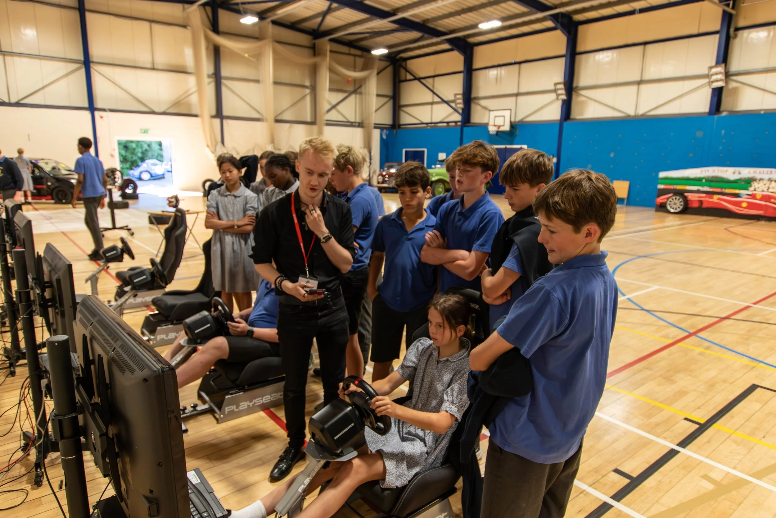 A group of children and Thomas Horton gathered in a gymnasium, watching someone race on a racing simulator