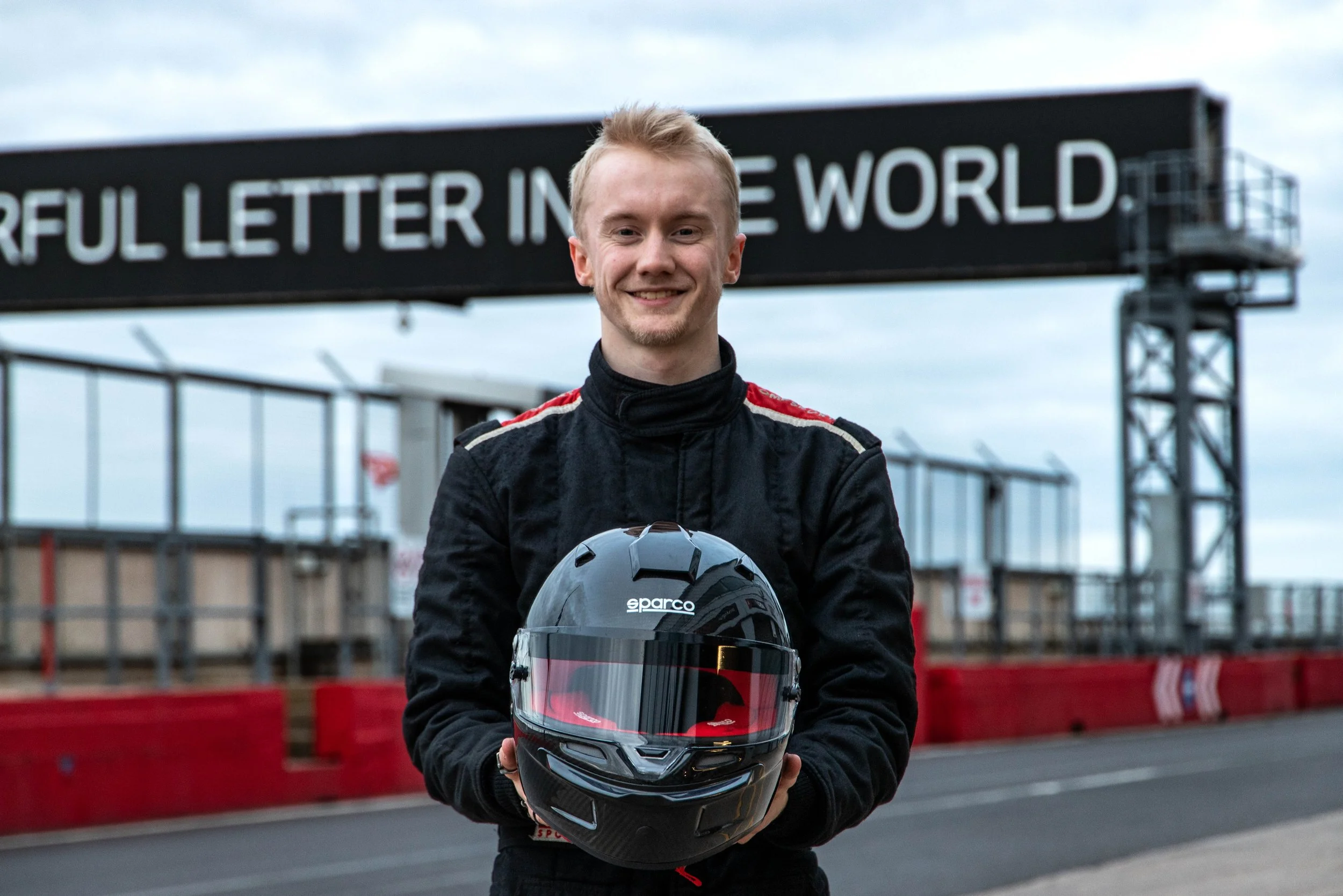Thomas Horton smiling while holding a racing helmet, standing on a race track with signs and fencing in the background.