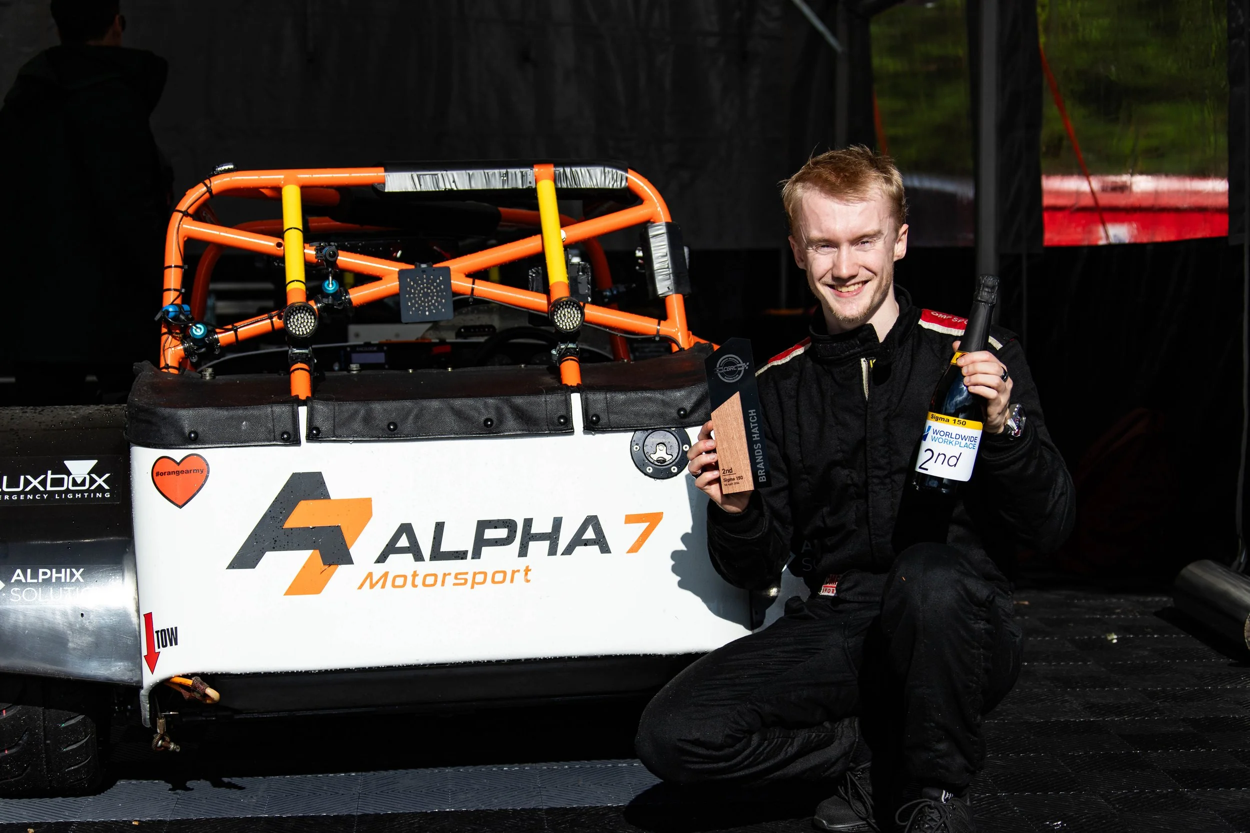 Thomas Horton in racing gear kneels next to his race car, smiling and holding a trophy and a bottle of champagne, with a black background and race track environment.