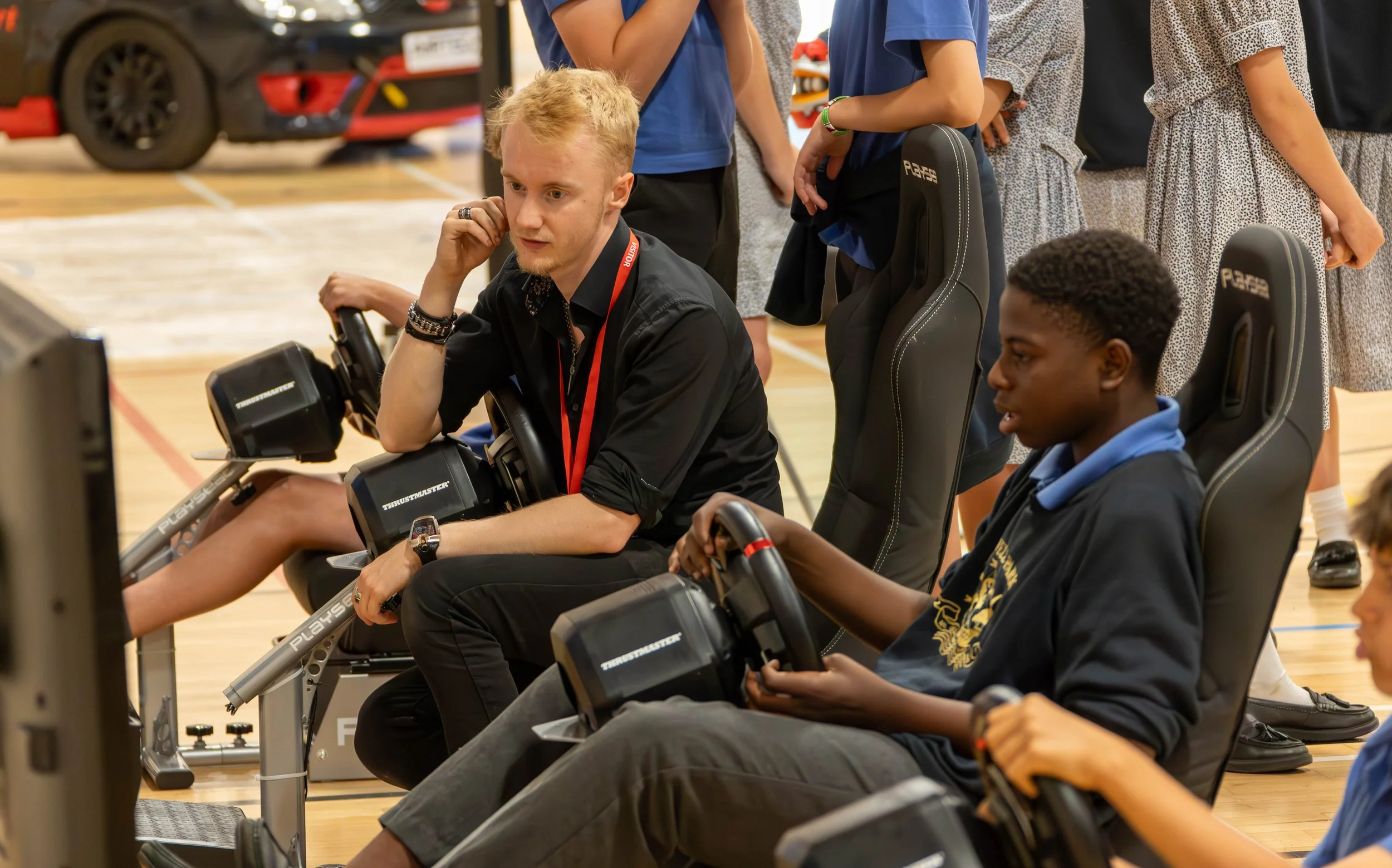 A group of children and Thomas Horton gathered in a gymnasium, watching someone race on a racing simulator
