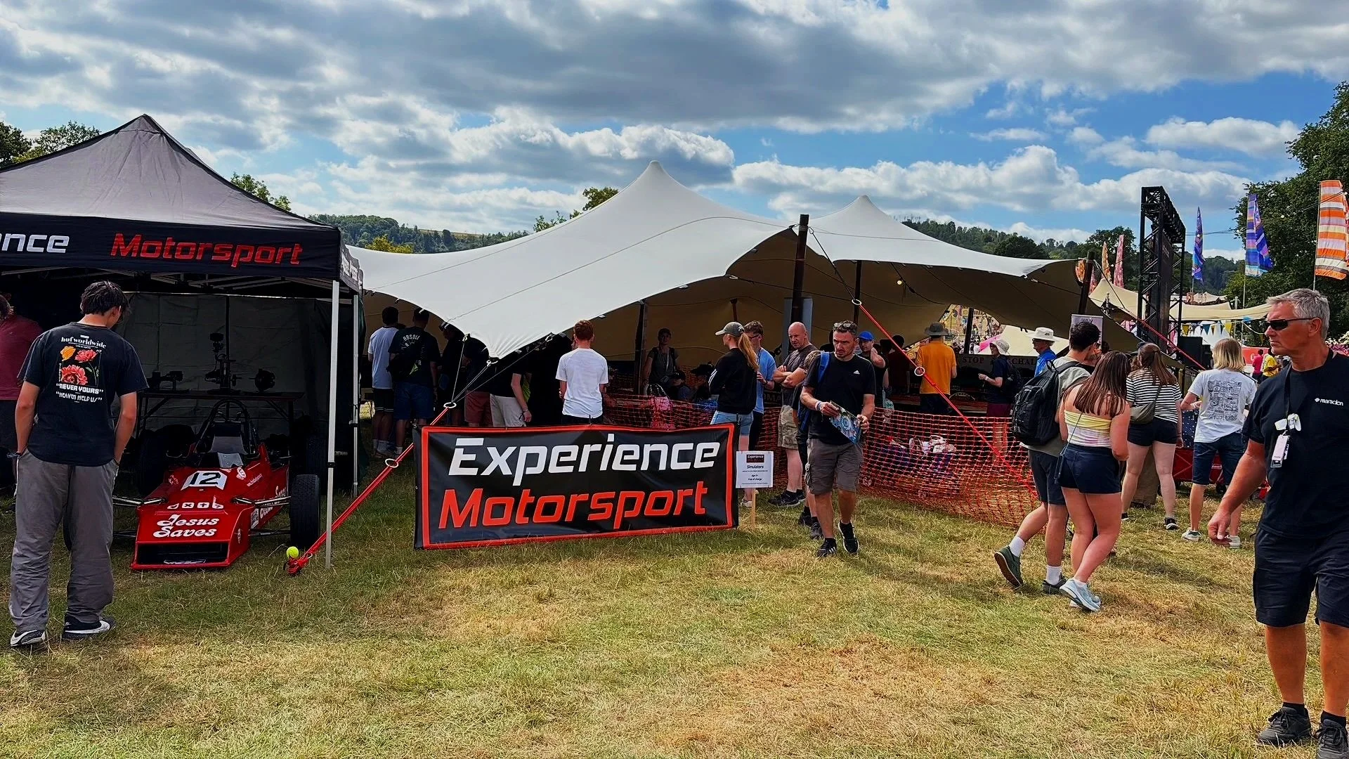 An outdoor event with a crowd of people gathered around tents and banners, including a black tent with red text that says 'Motorsport' and a large sign that reads 'Experience Motorsport.'