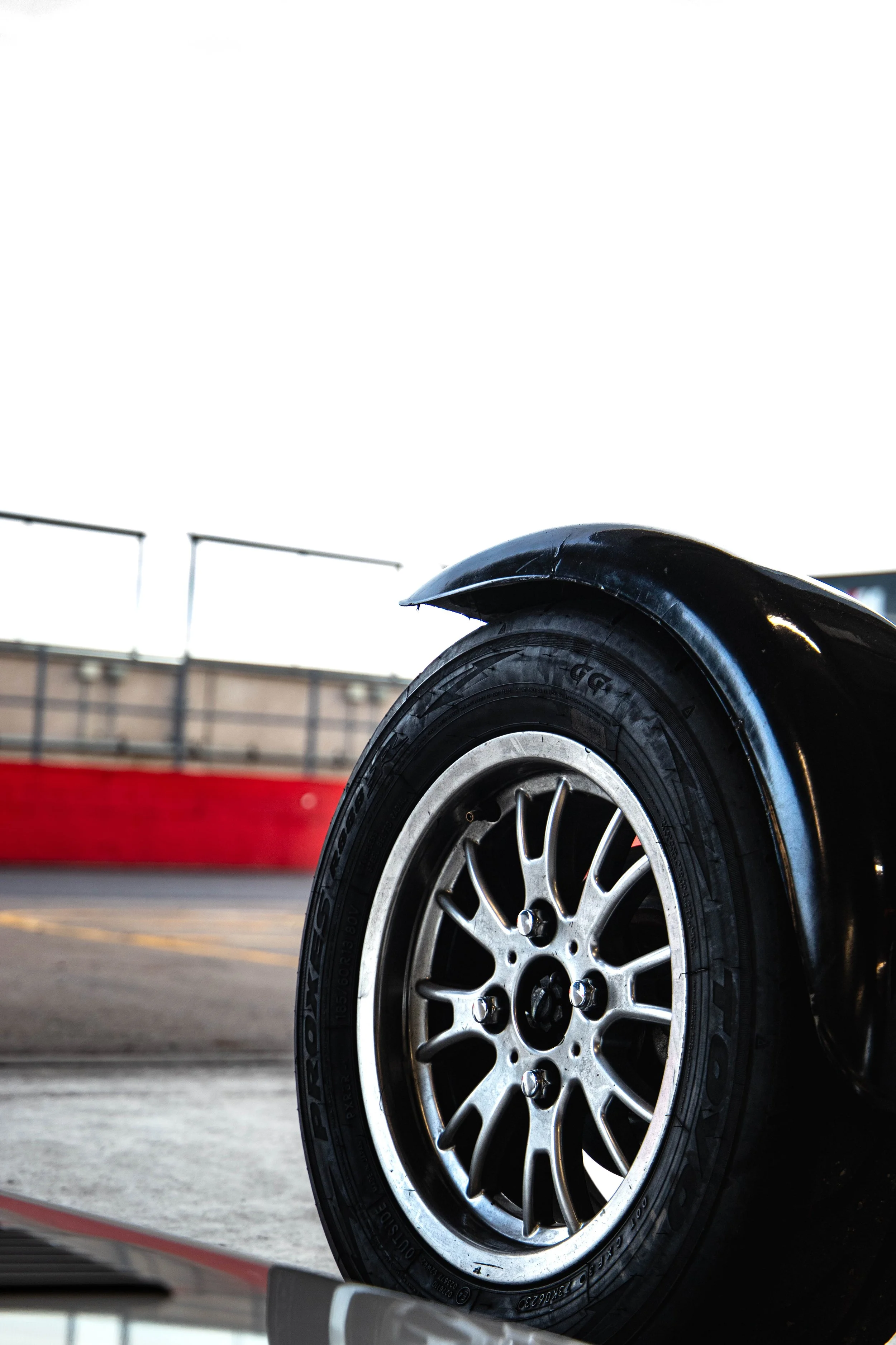 Close-up of a car tire and wheel, part of a black vehicle, with a building and fence in the background