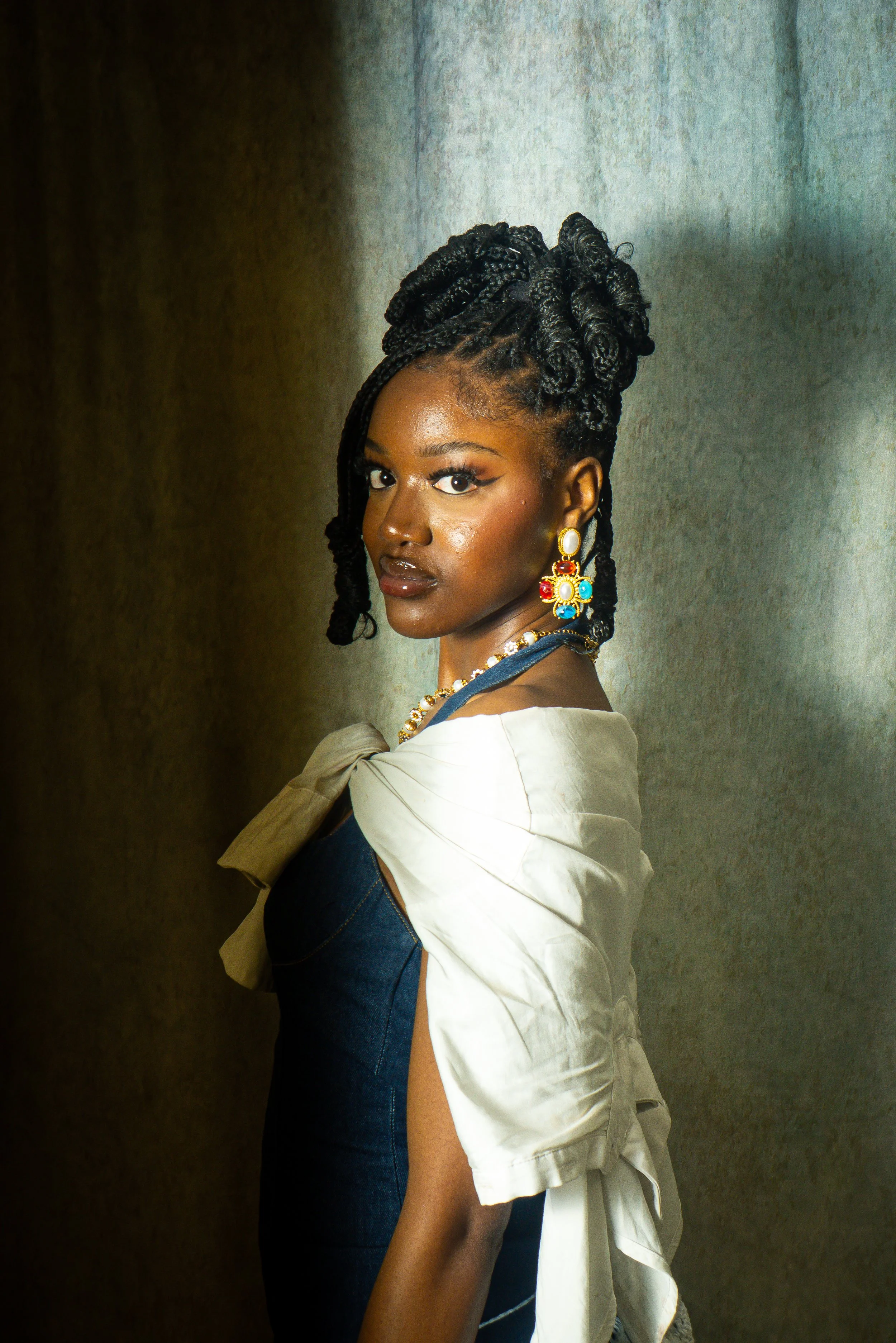 A woman with braided hair styled in an updo, wearing statement earrings and a necklace, dressed in a dark dress with a cream-colored shawl draped over her shoulders, standing against a textured, metallic background.