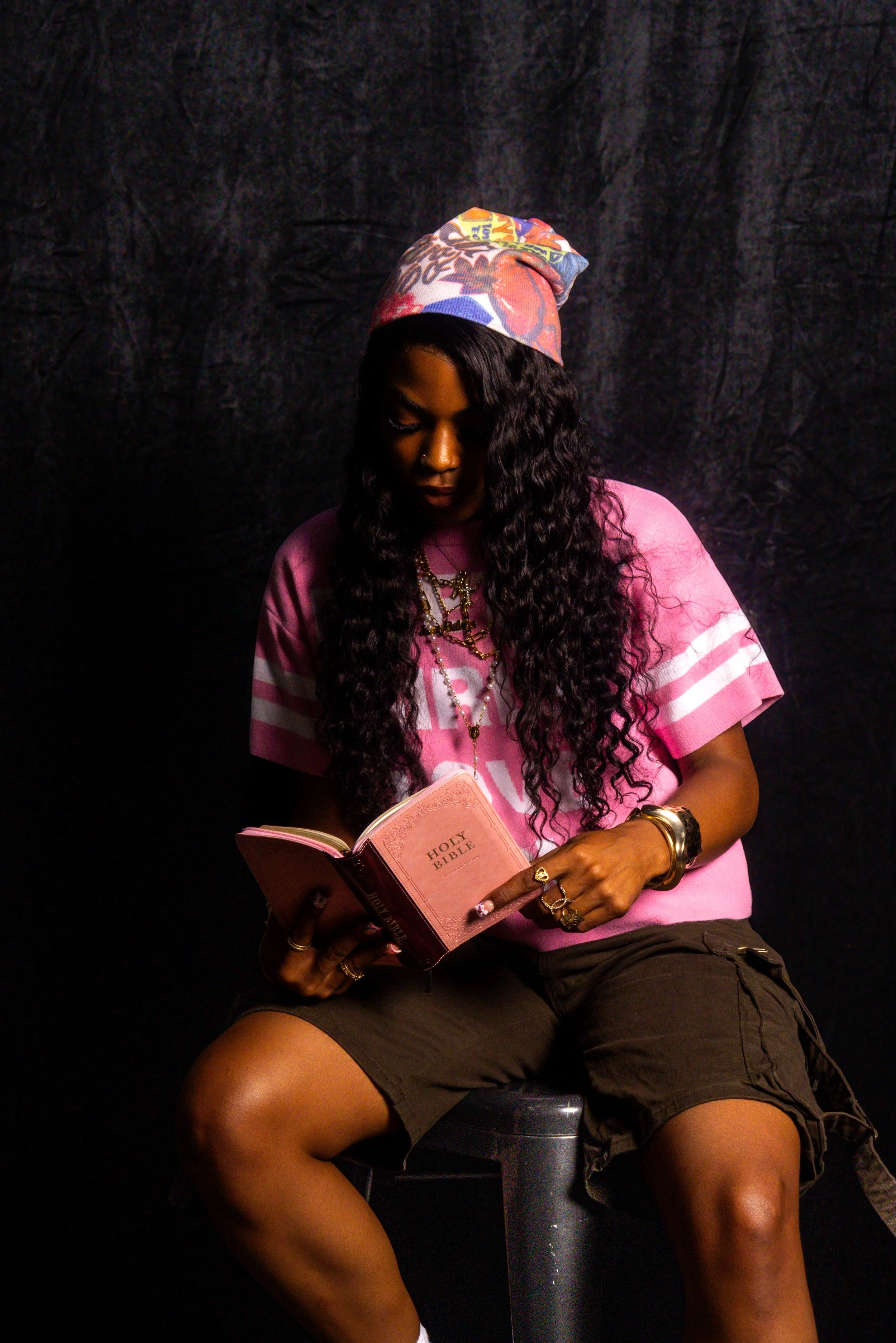A woman with long curly hair, wearing a pink t-shirt and a colorful beanie, sitting on a stool and reading a pink 'Holy Bible,' against a dark background.