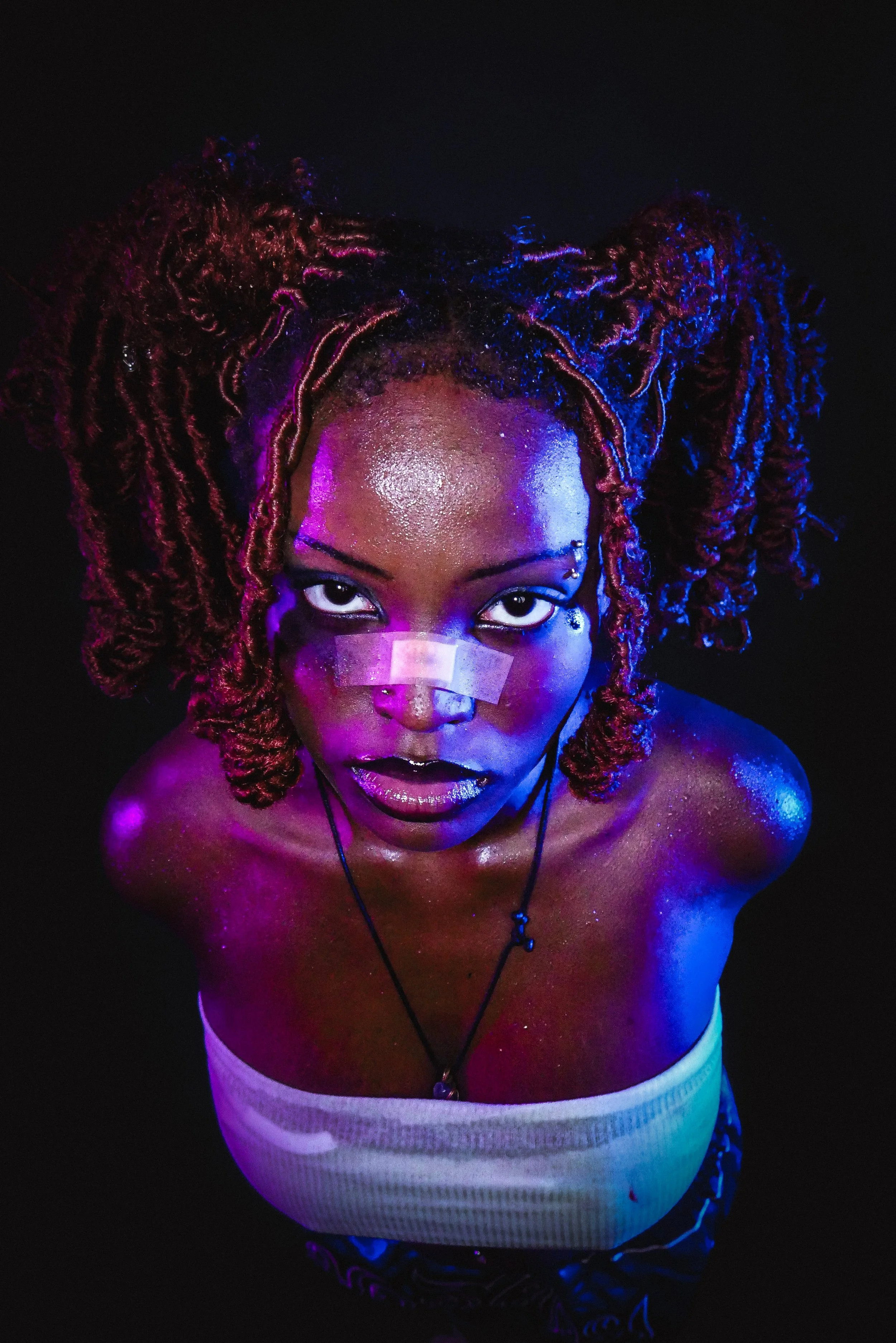 A woman with dreadlocks styled in two large puffs, adorned with glitter and neon paint, looking directly at the camera against a black background.
