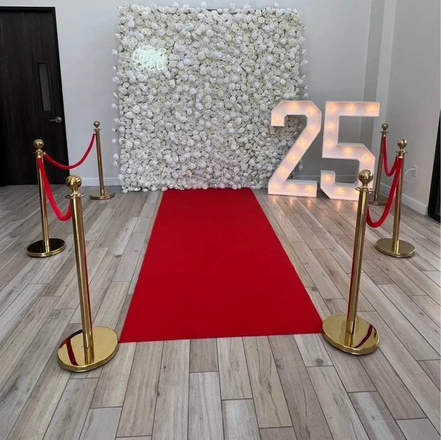 Red carpet flanked by red velvet ropes on gold stanchions leads to a backdrop of white flowers and illuminated number "25" at an event or celebration. For Houston and surrounding areas. Event rentals.