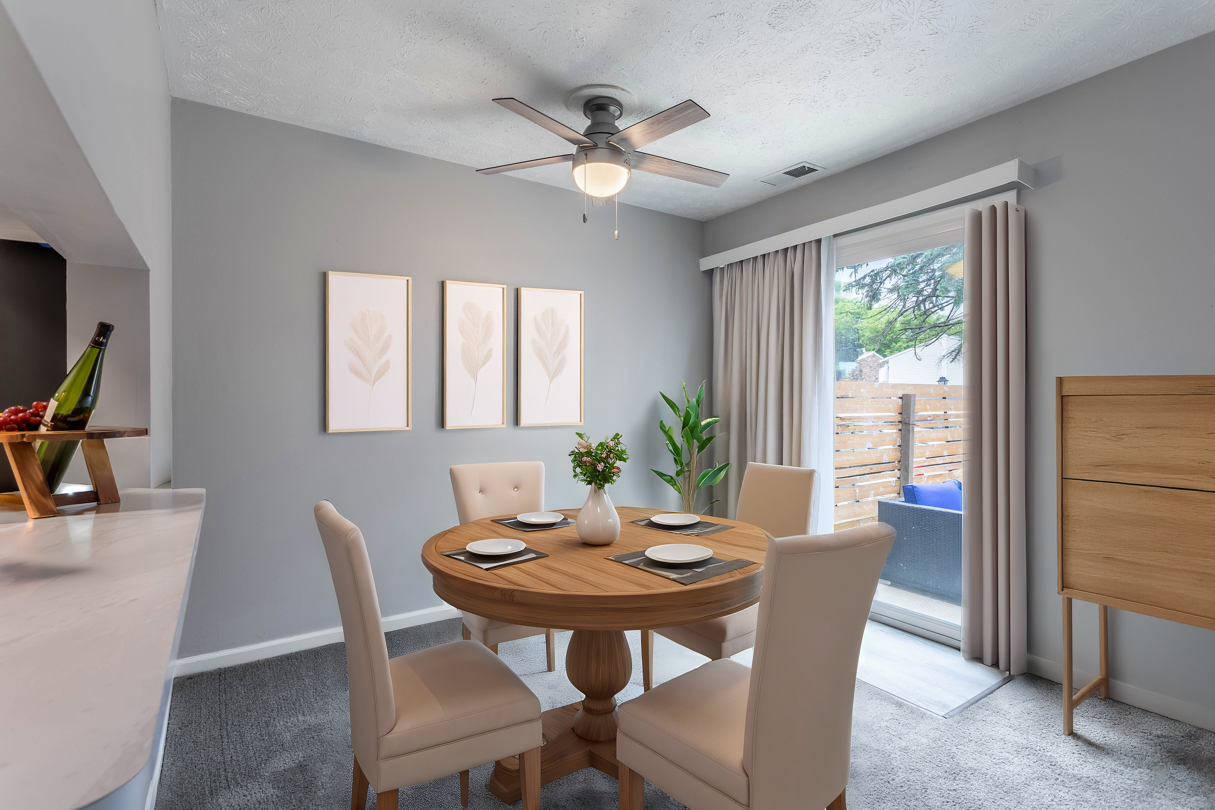 A dining area with a round wooden table set with four plates and napkins, cream-colored chairs, a vase with flowers, and a large window with curtains. There is a ceiling fan and artwork on the wall.