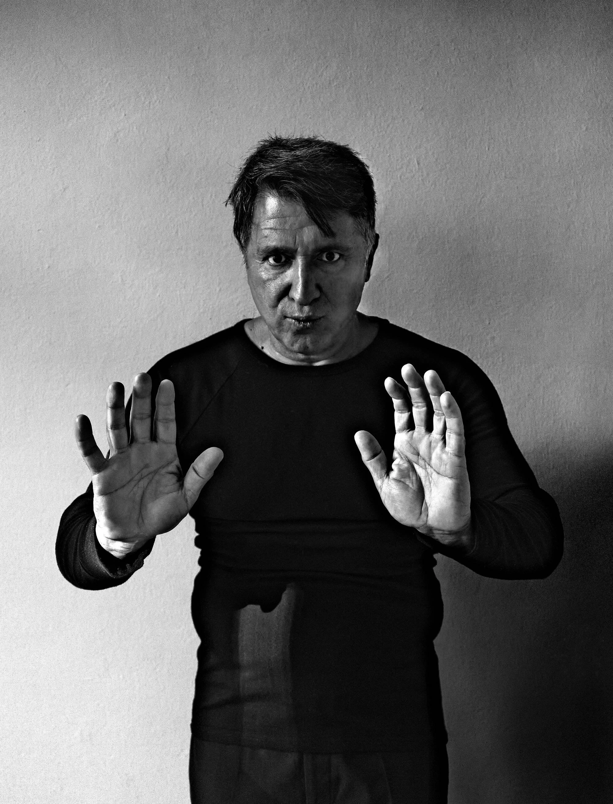 A man with dark hair and intense stare holding up both hands with palms facing forward, in a black shirt against a plain wall.