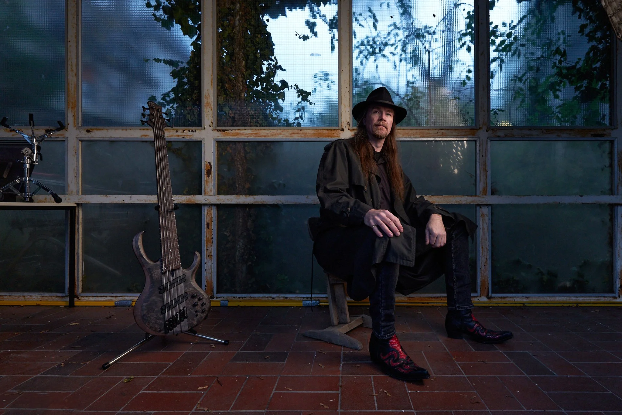 A man with long hair, a beard, and wearing a black hat and black clothing sitting on a bench outside. There is a guitar on a stand and a drum set nearby, and behind him is a rusty metal frame with large windows showing trees and the sky.
