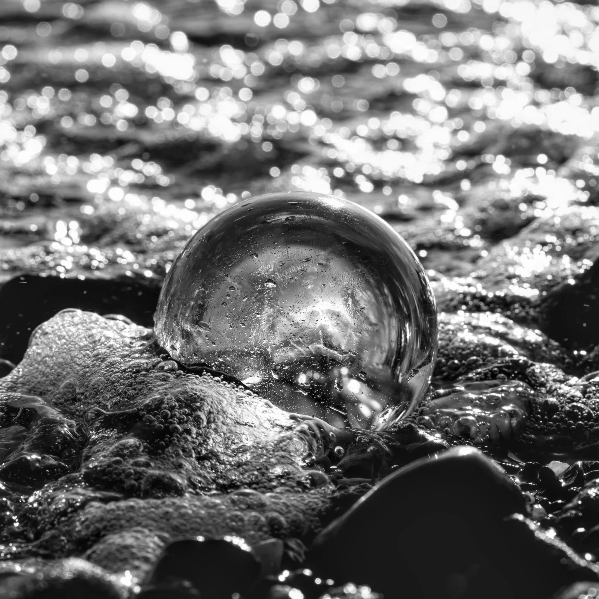 A clear glass sphere on a pebbled shoreline with water and sunlight reflections around it in black and white.