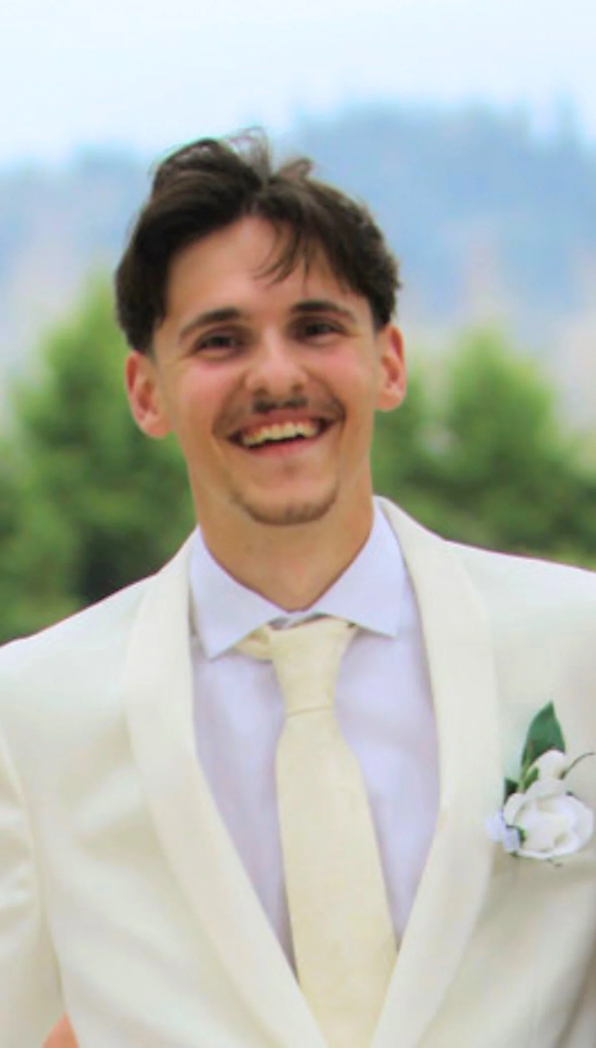 A young man in a white suit and cream tie, smiling outdoors with blurred green trees and cloudy sky in the background.