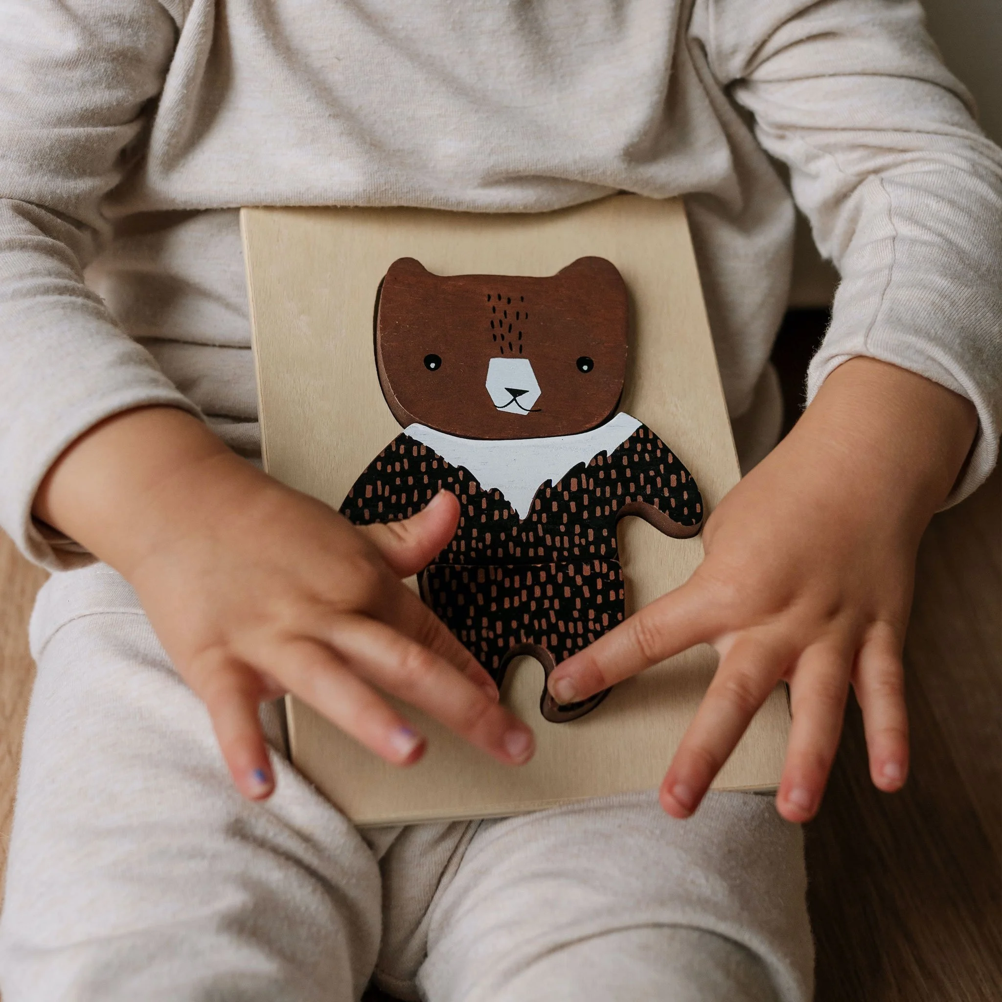 Child holding a wooden bear puzzle piece on their lap, sitting on a beige chair.