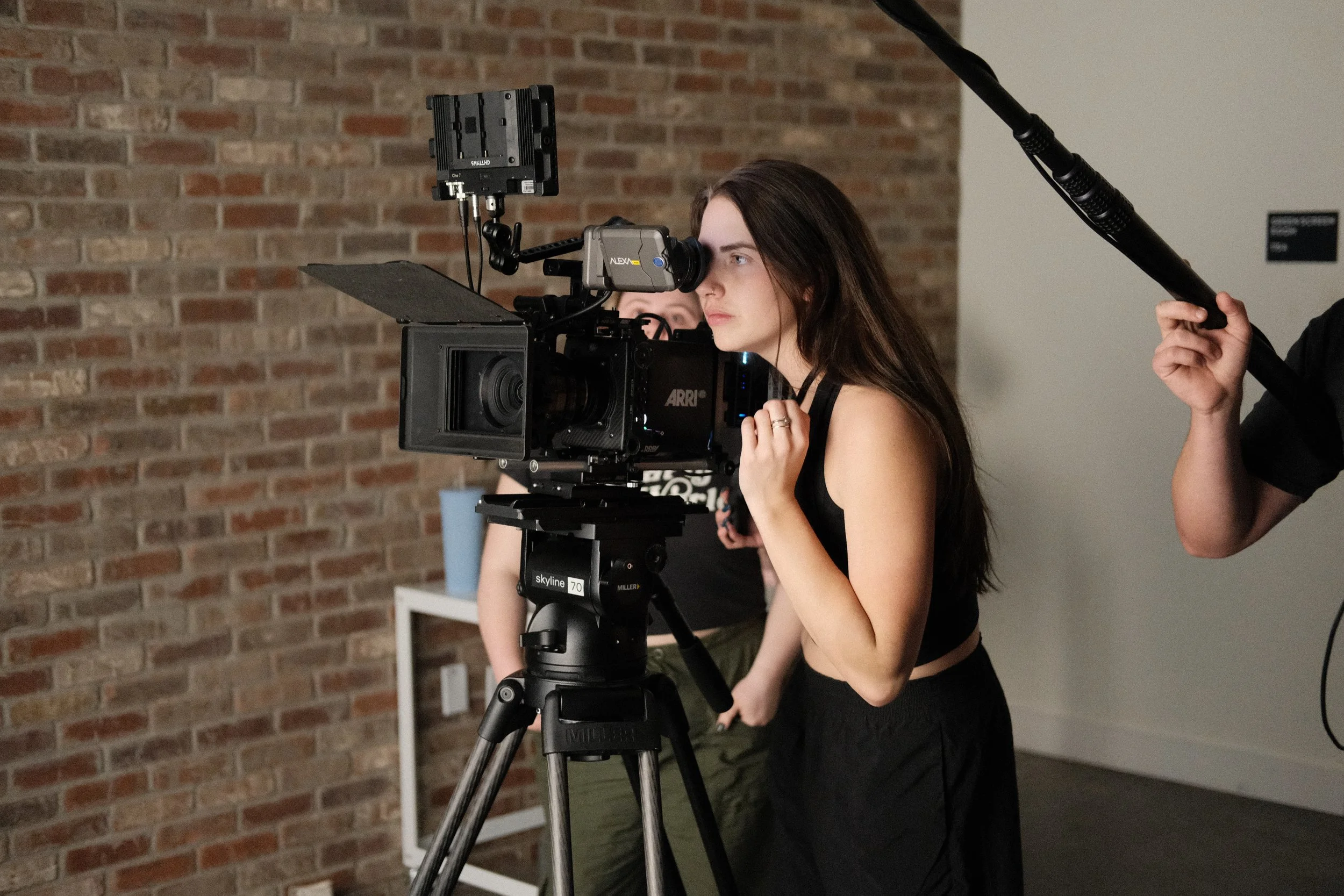 A young woman with long dark hair looking through a professional camera mounted on a tripod, on a set with a brick wall background.