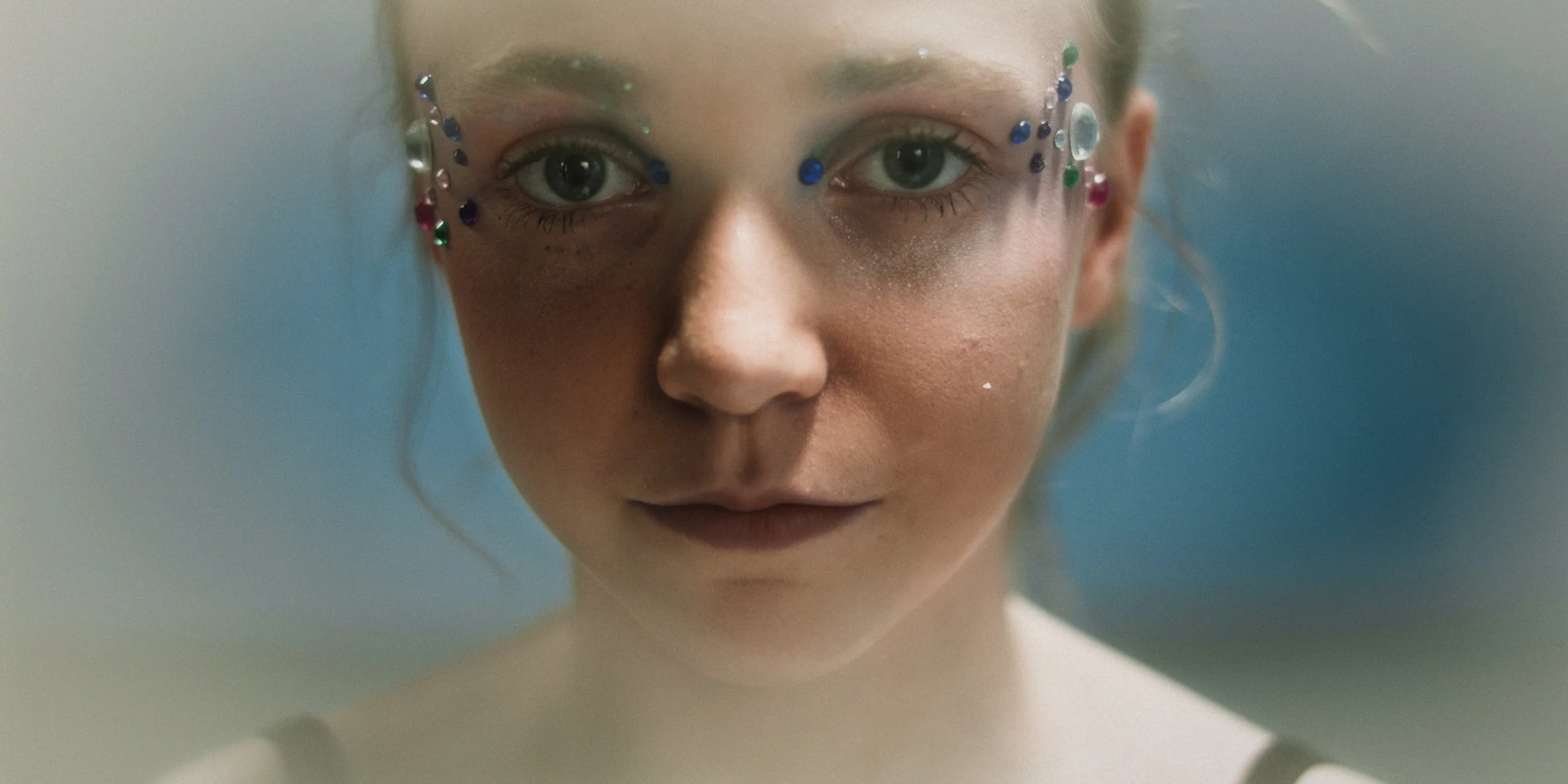 A close-up of a woman's face with light makeup, blue eyes, and decorated with colorful rhinestones around her eyes.