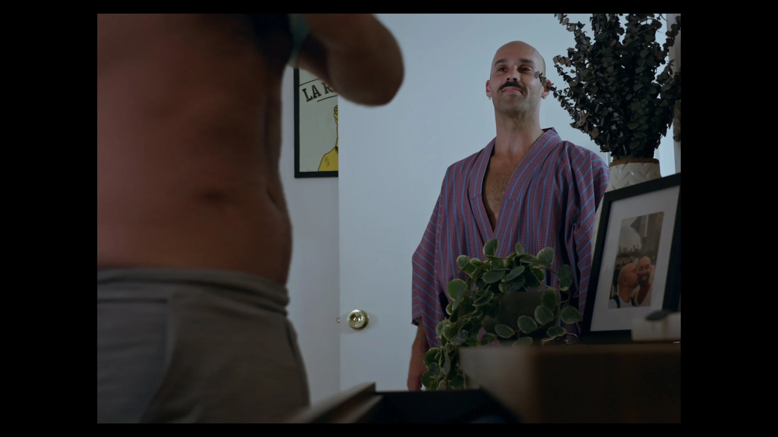 A man with a mustache standing in a doorway, wearing a striped robe, with a neutral expression. In the foreground, part of a person's midsection is visible, with a dresser and framed photos and plants on top.