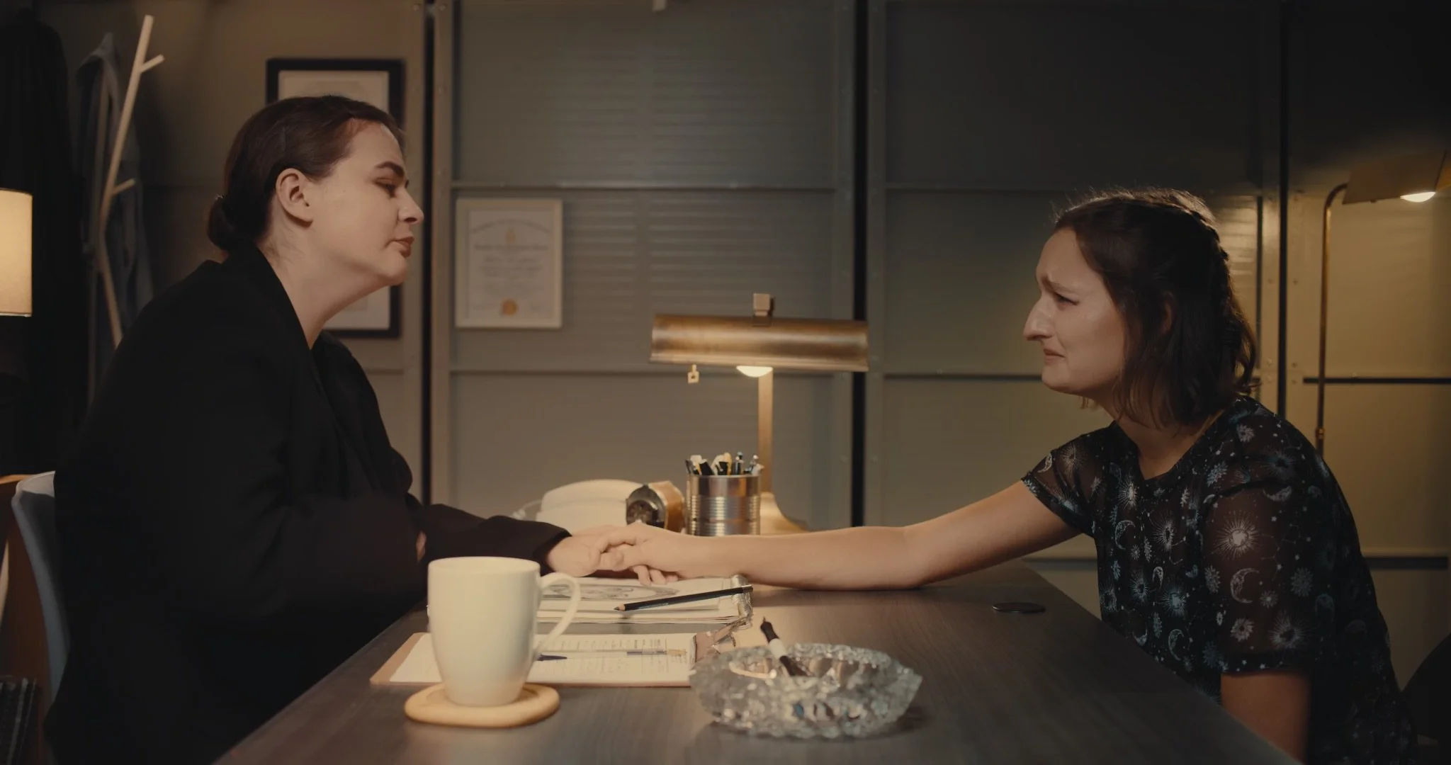 Two women sitting at a table, holding hands and sharing an emotional moment, in an office/investigation room.